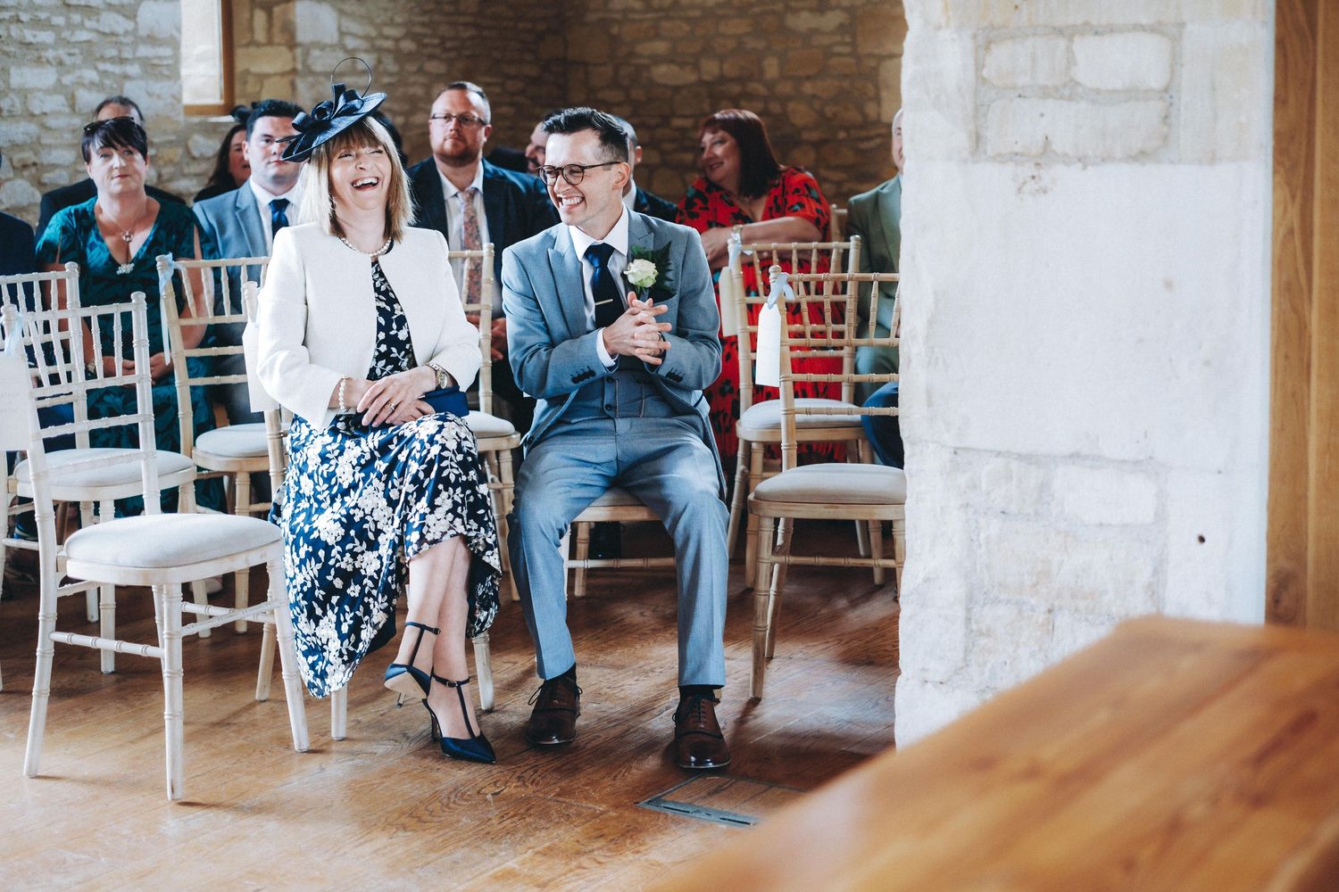 Wedding guests seated on white chairs in a rustic brick room look at their phones during the ceremony.