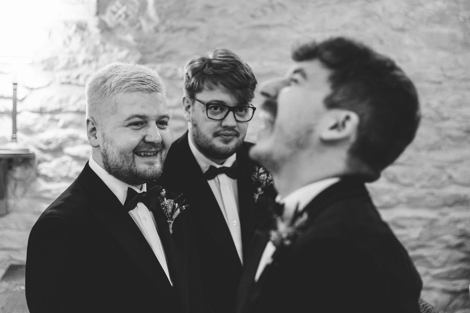 Three groomsmen share a candid laughing moment in black tuxedos against a stone wall background.