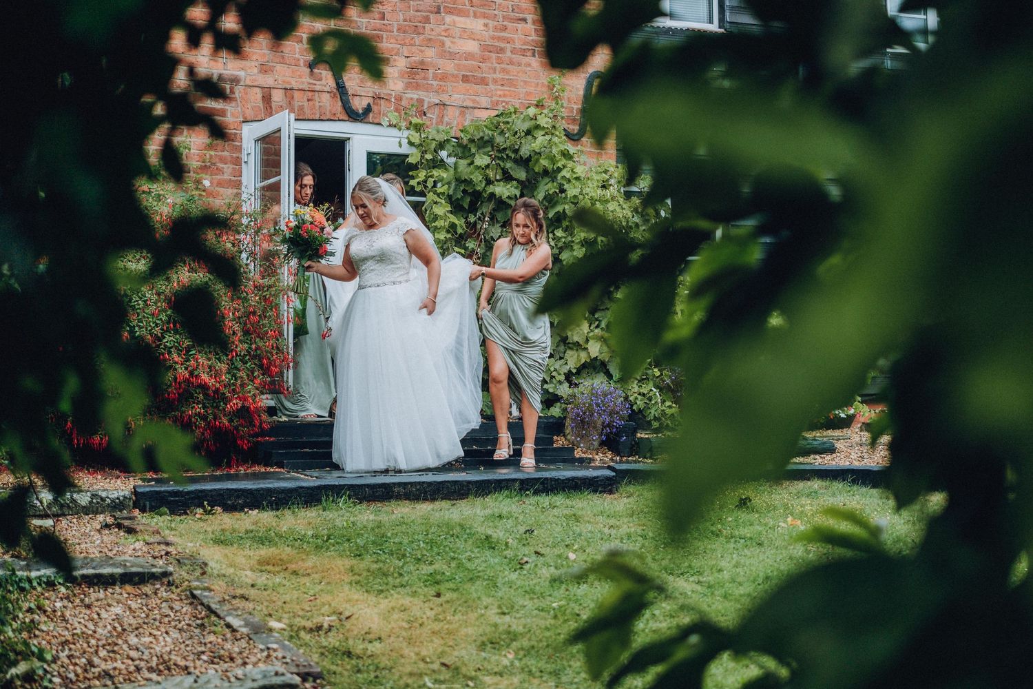 Wedding guests gather outdoors in a garden setting with lush greenery and brick walls.