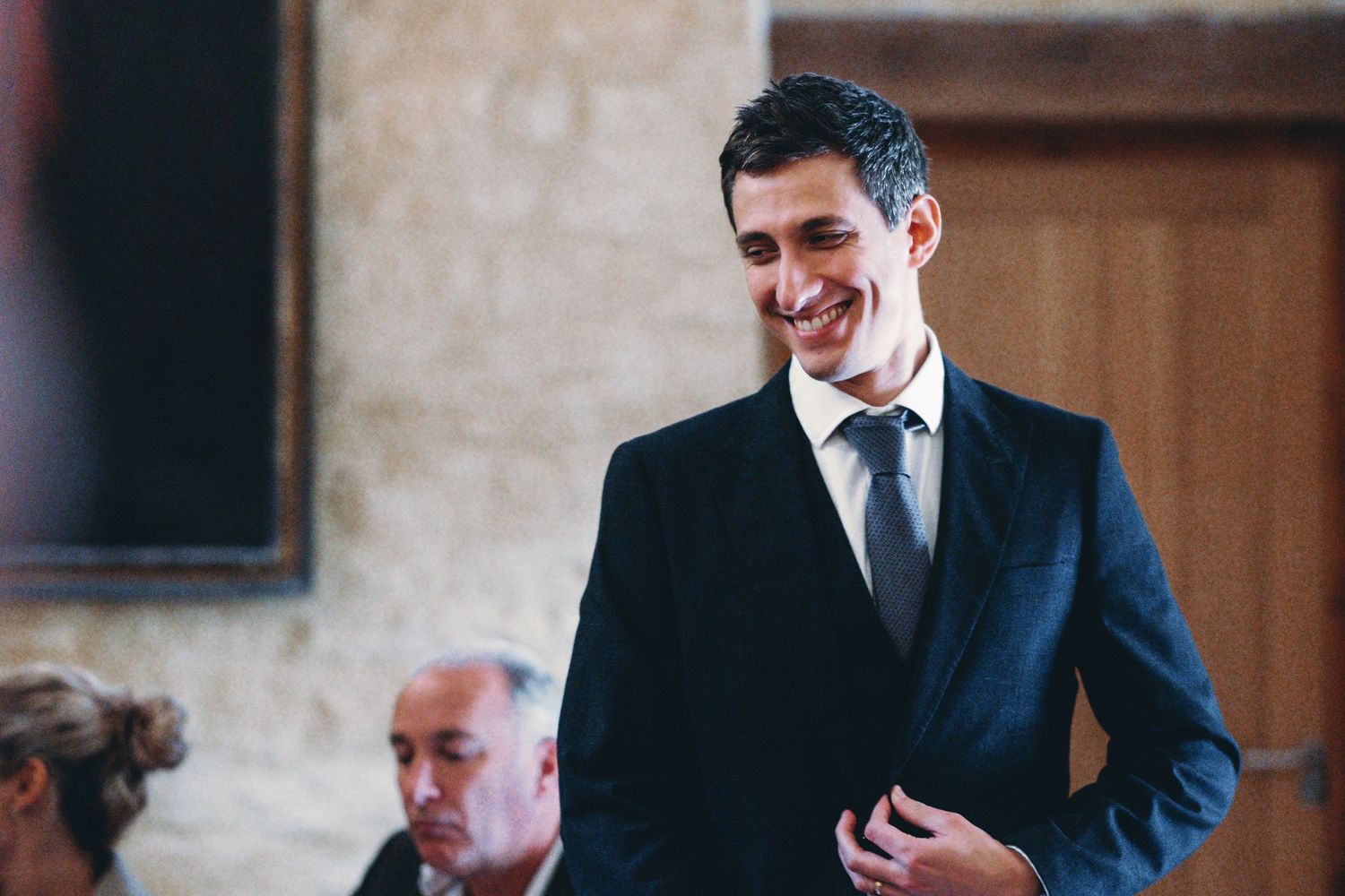 A person in a navy suit smiles warmly during a formal indoor gathering.