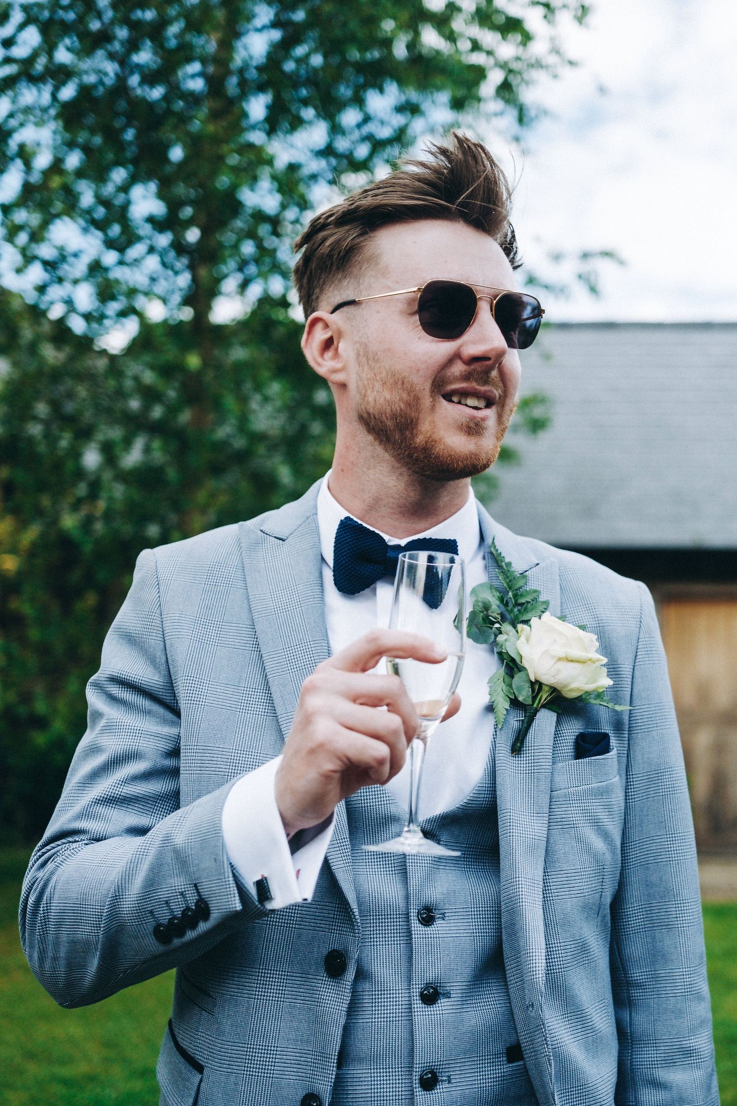 Wedding guest in light grey suit and sunglasses holds champagne glass while smiling outdoors.