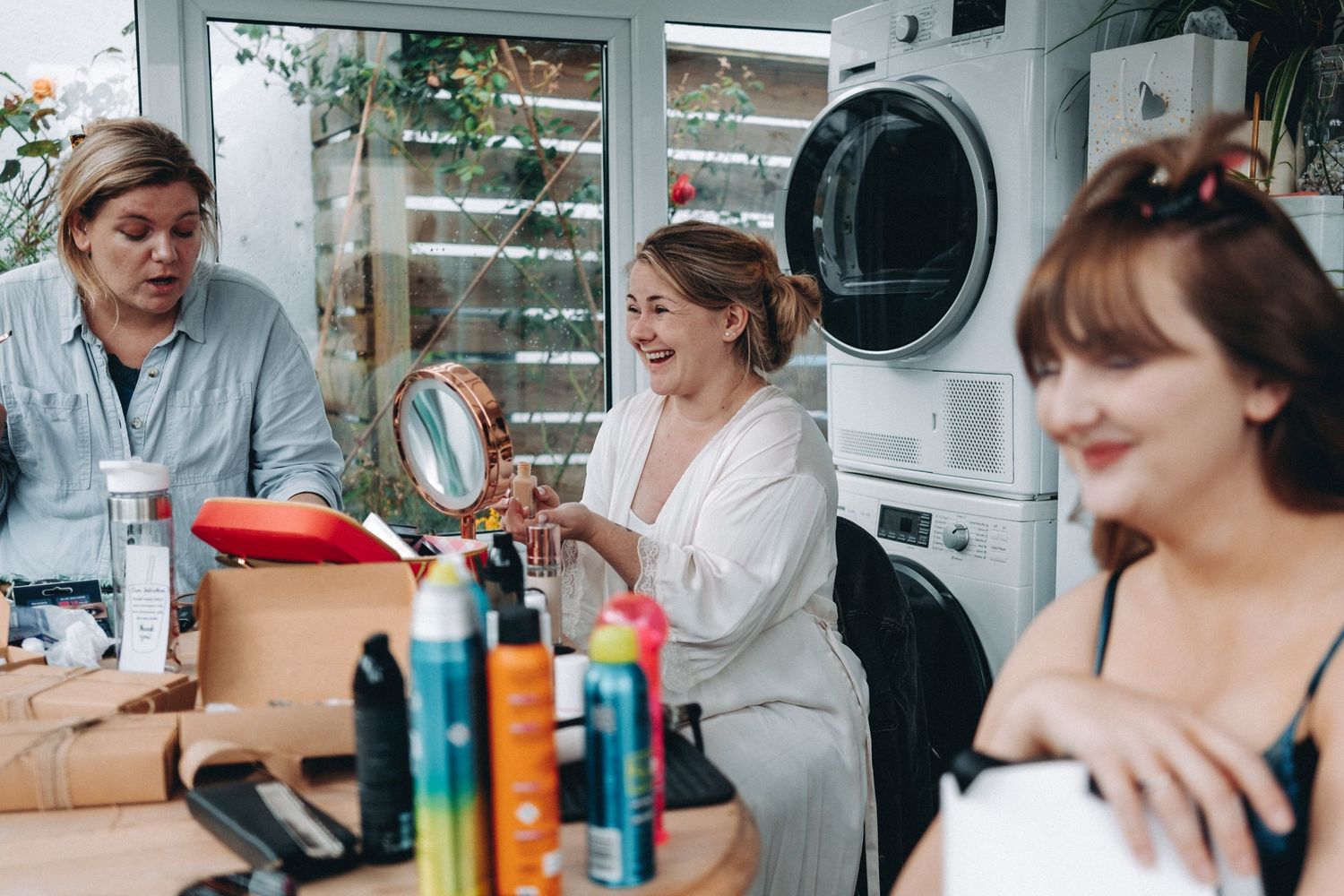 Bridal group relaxes in casual setting near laundry room with drinks and snacks while socialising and applying makeup.