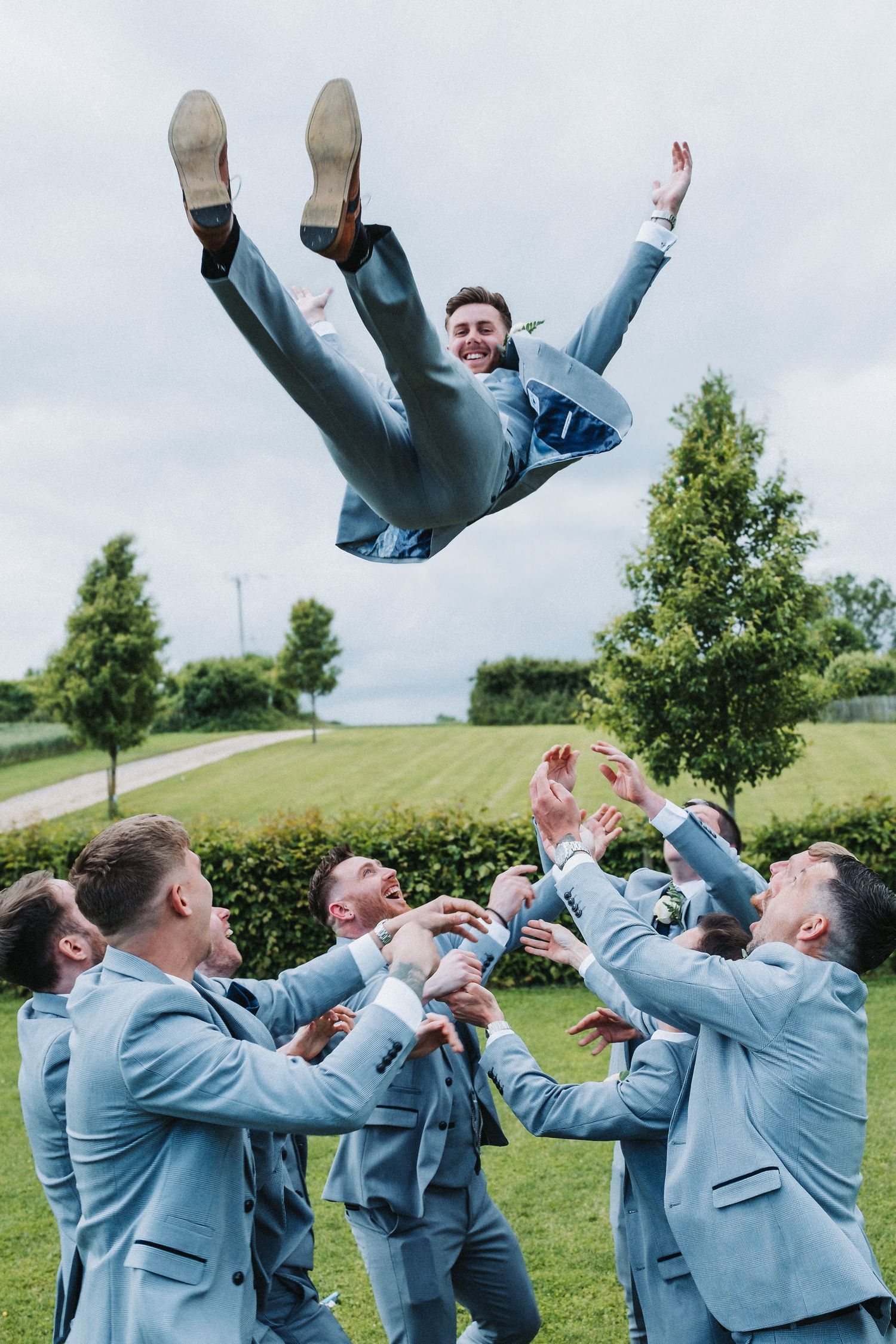Groomsmen in light blue suits toss someone in the air on grassy lawn.