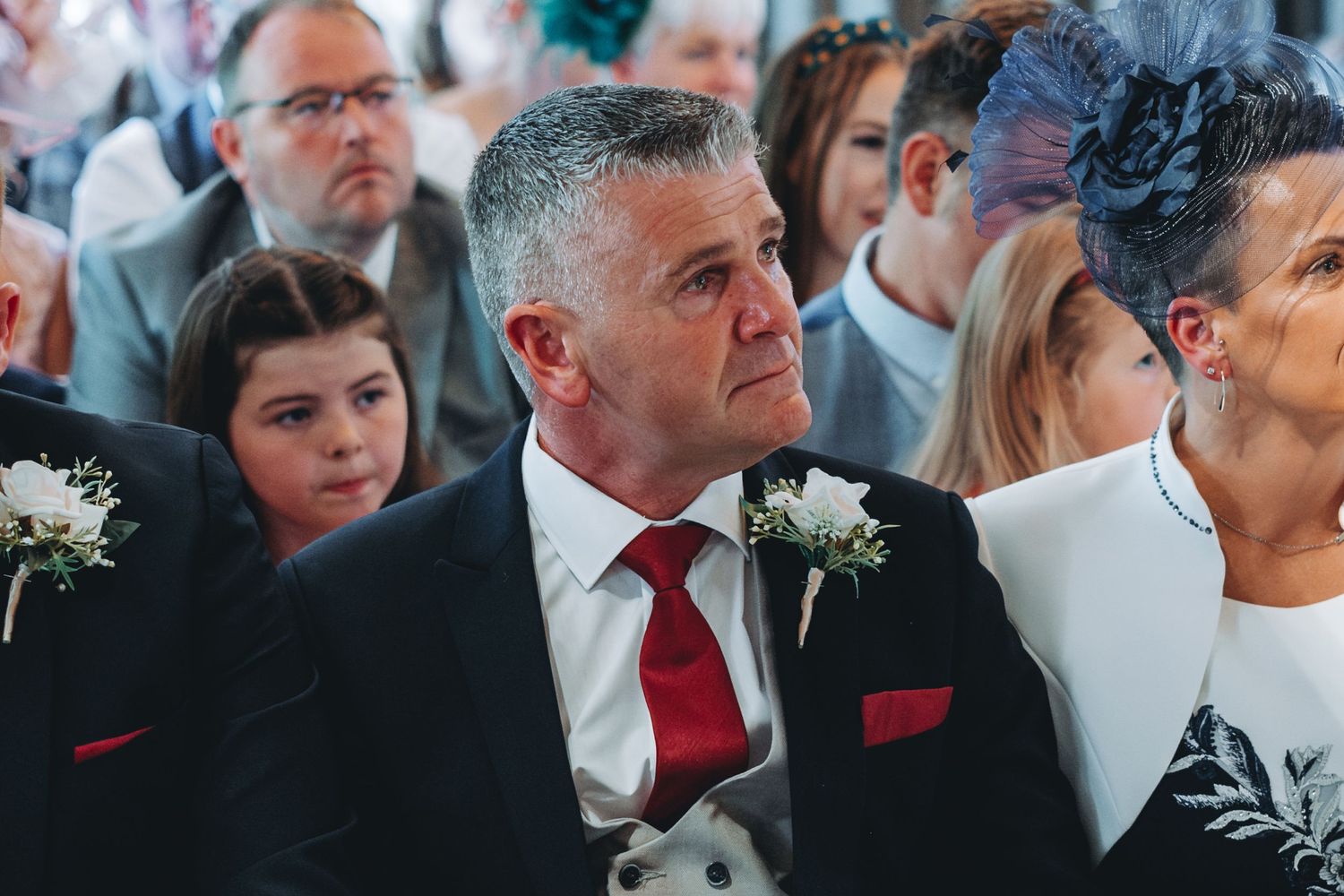 Wedding guests seated in formal attire during ceremony with brick wall backdrop.