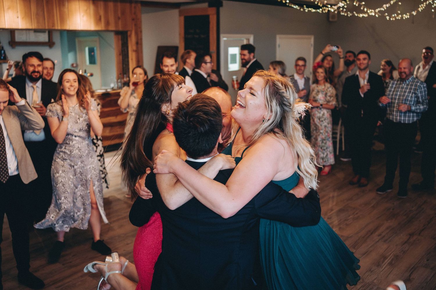 Party guests dance and celebrate together on a wooden dance floor under string lights at a wedding event.