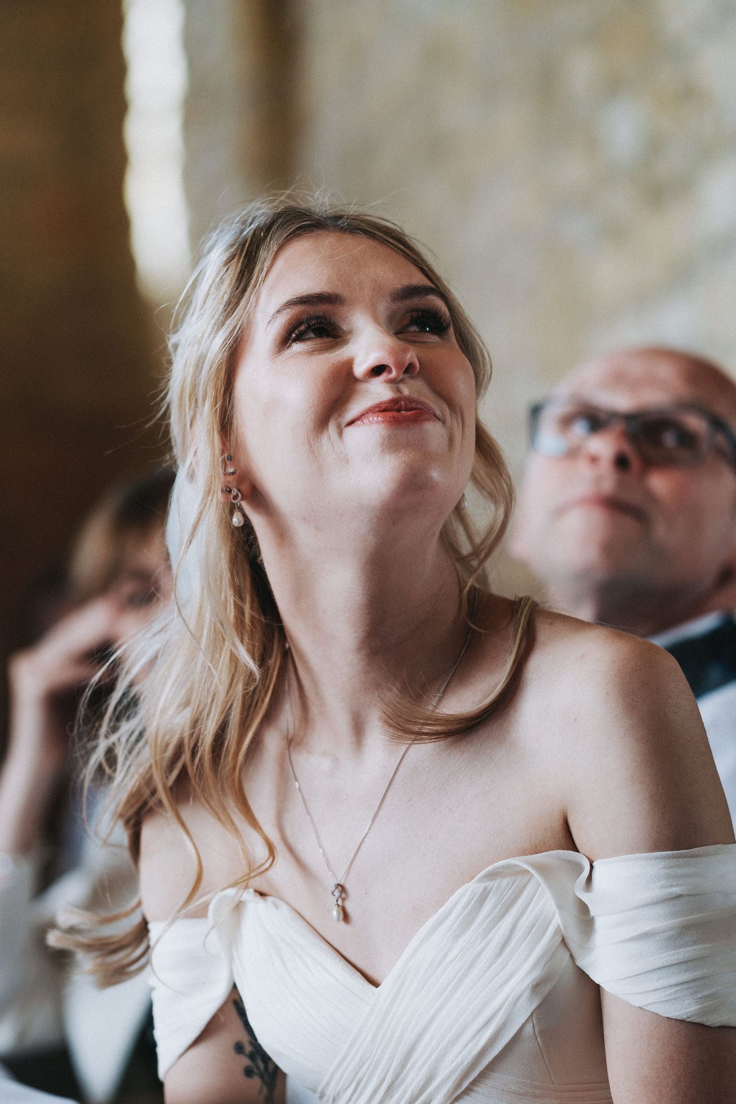 A bride in an off-shoulder white wedding dress smiles joyfully while looking up during an indoor celebration.