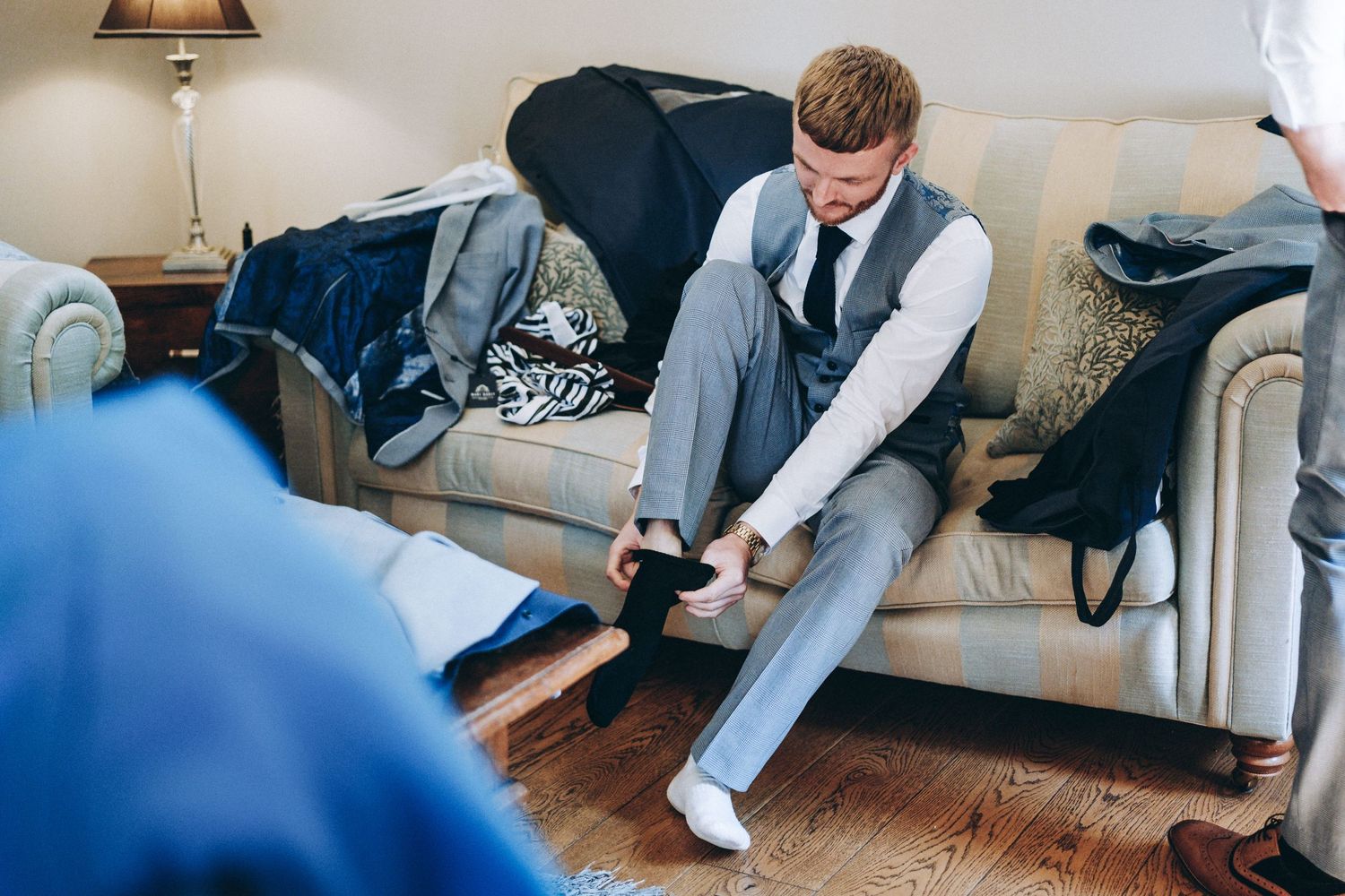 A person in formal gray attire adjusts their dress shoes while sitting on a couch during preparation.