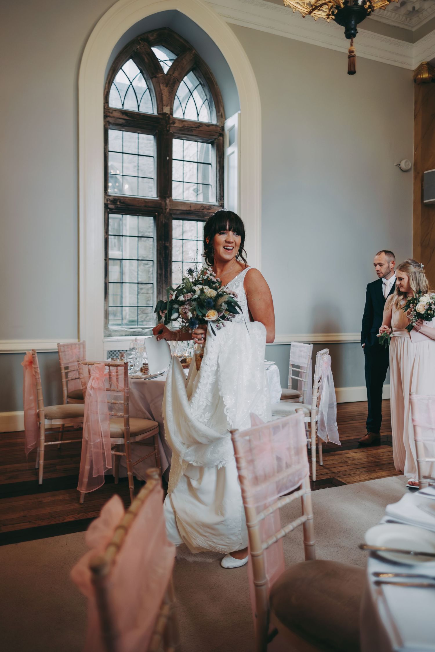 A bride in a white gown stands in an elegant room with arched Gothic windows and pink chair decor.