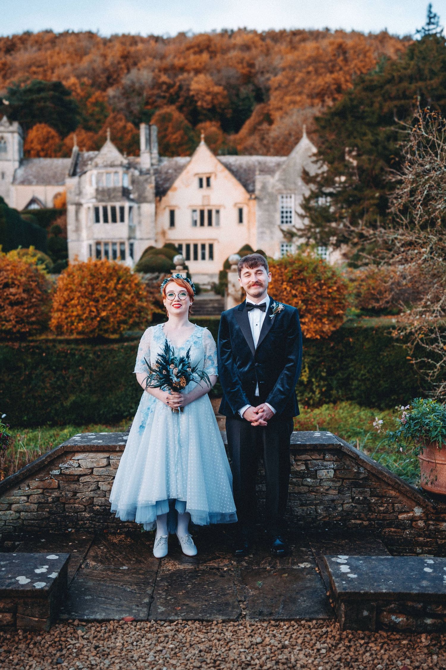 A couple stands on stone steps in front of a grand historic Owlpen Manor with autumn autumnal foliage in the background.