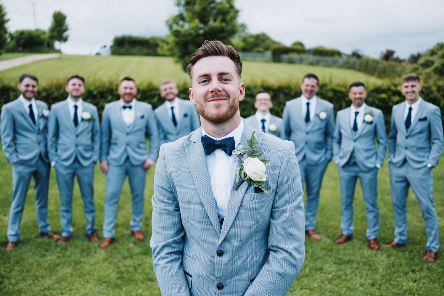 Wedding party wearing matching light blue suits poses together on green grass against scenic countryside backdrop.