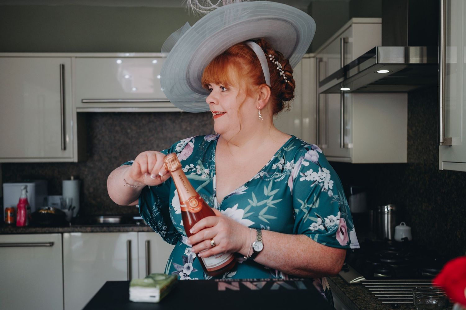 mother of the bride in a teal floral dress and white hat opens a bottle of champagne.