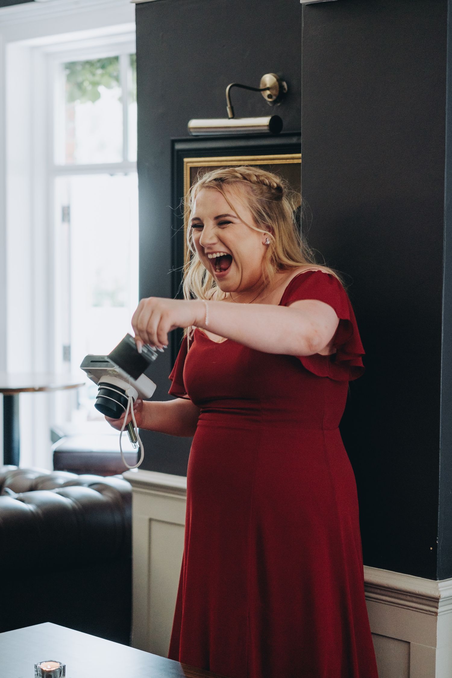 Someone in a flowing red dress laughs joyfully while printing a polaroid in a bright wedding venue setting.