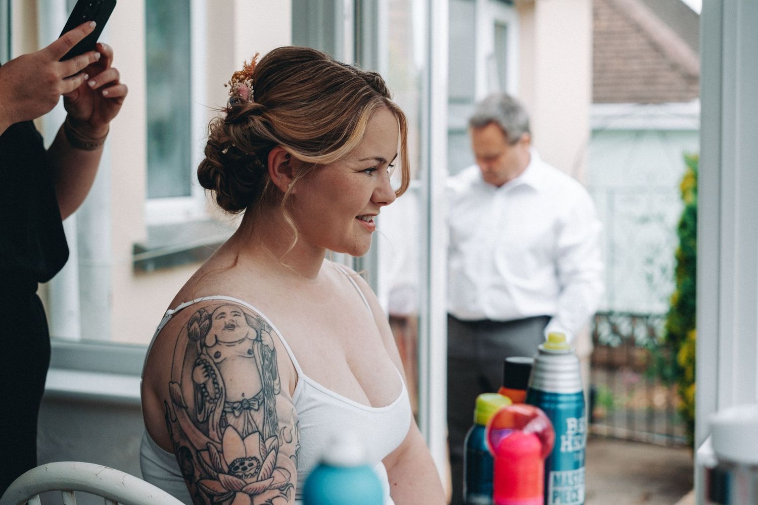A bride with detailed arm tattoos gets ready while dad stands in the background on a porch.