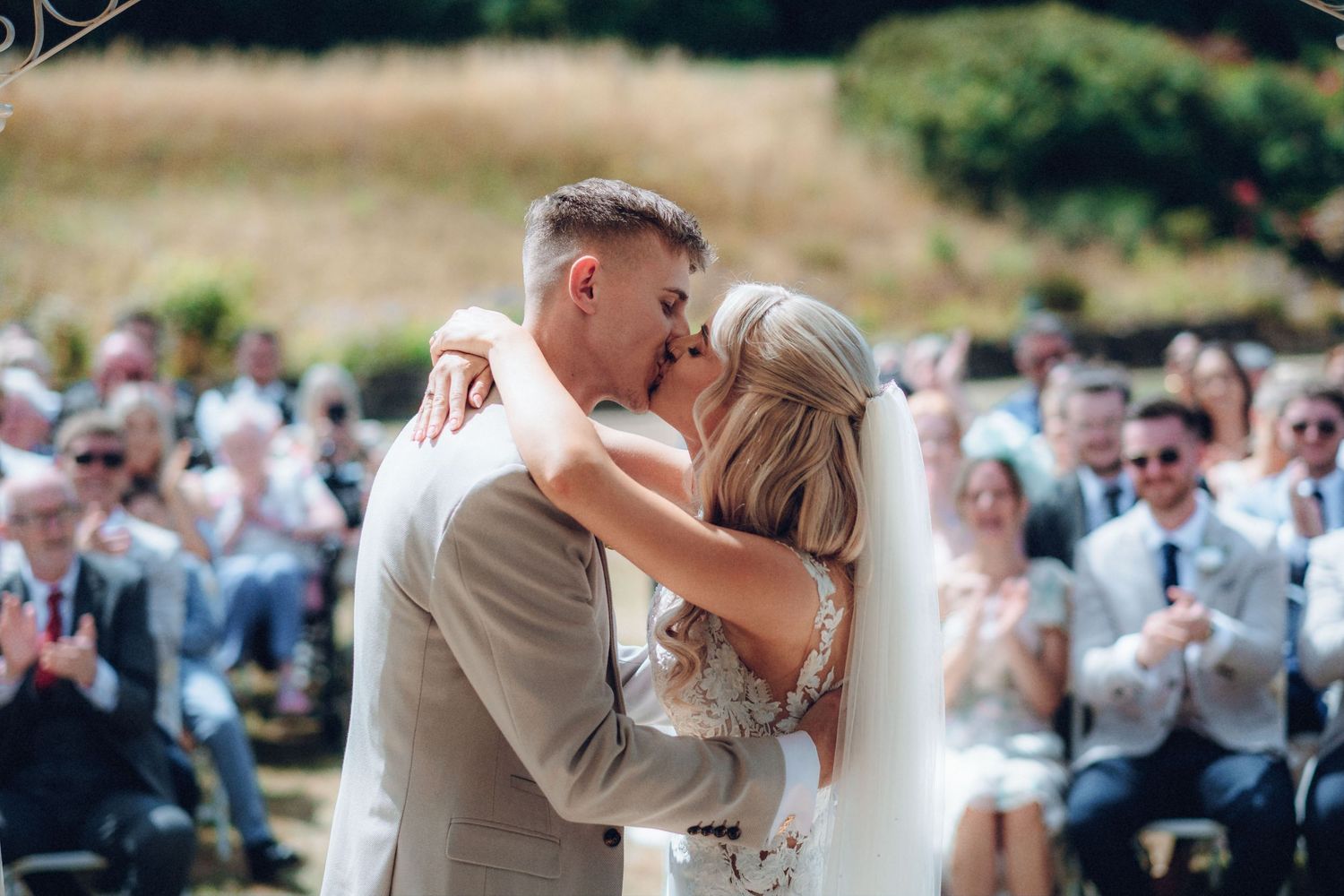 Newlyweds share their first kiss at outdoor ceremony as guests applaud.