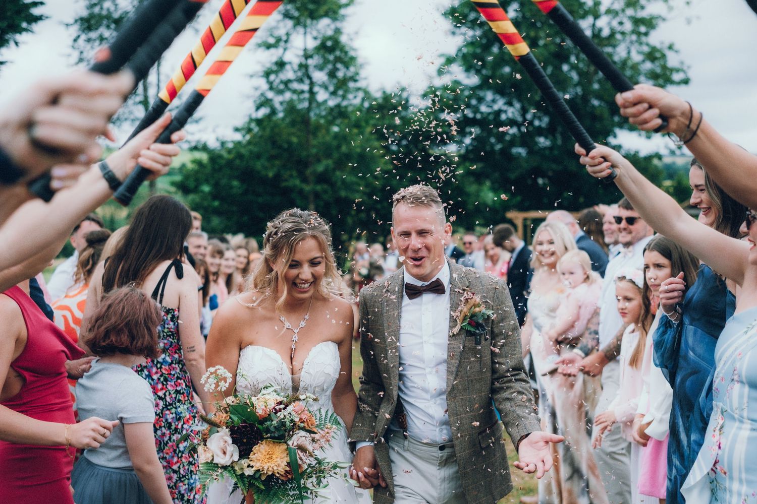 Wedding couple walks through guest tunnel with raised sparklers during outdoor celebration.