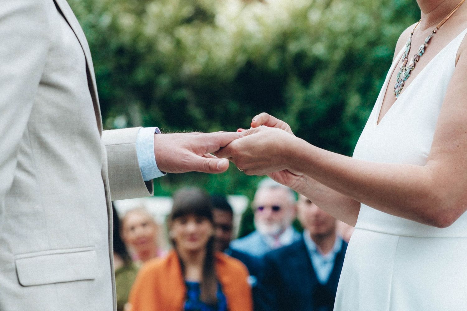 Close-up of wedding rings being exchanged during an outdoor ceremony with guests in background.