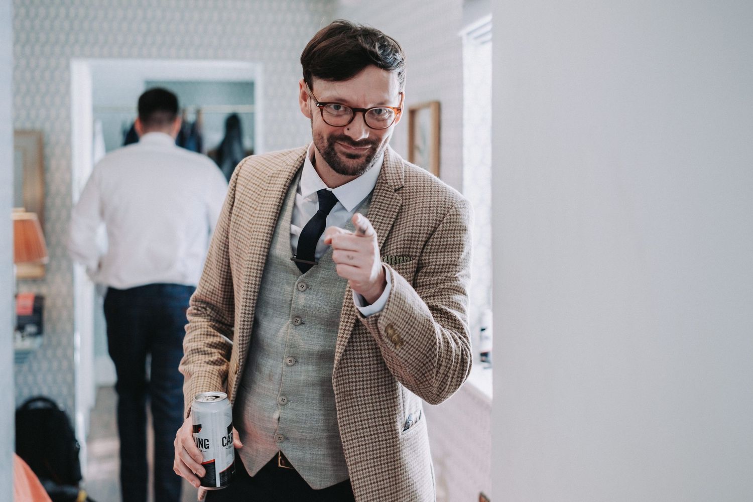 A person in a beige blazer and vest smiles while walking through a hallway.