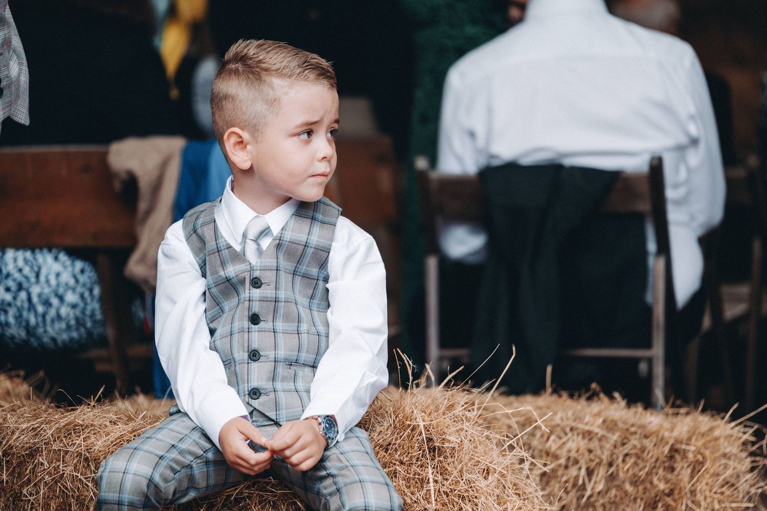 Young child in stylish gray plaid vest and dress pants sits thoughtfully on hay bale during formal event.