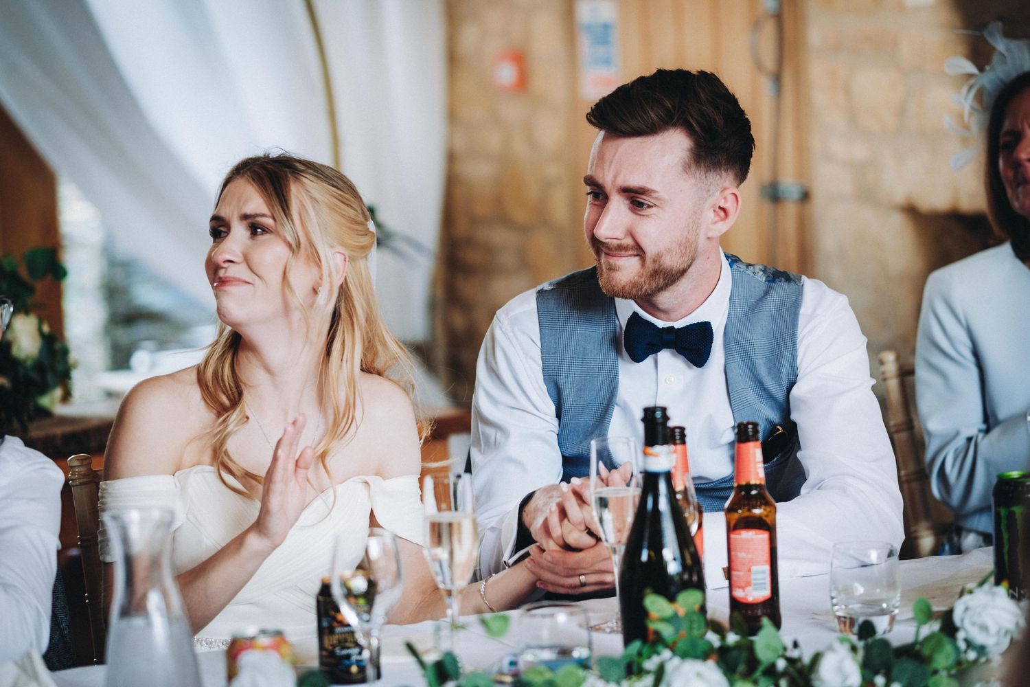Newlyweds sit at an elegant reception table with wine bottles and floral decorations during wedding dinner celebration.
