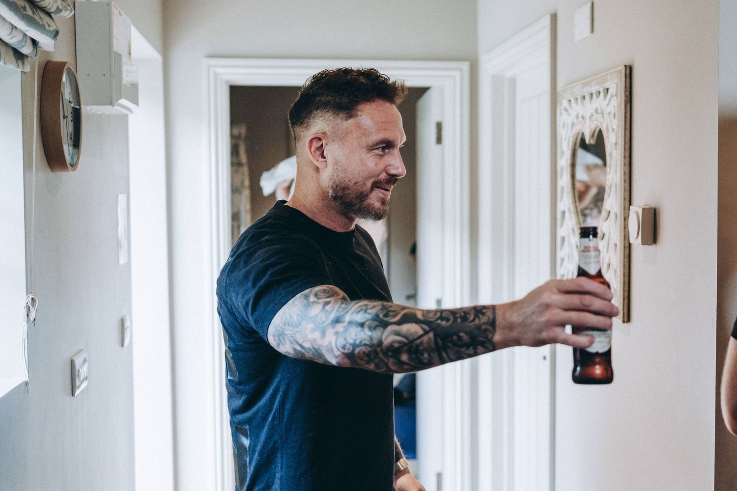 Person with tattooed arm reaches for bottle in hallway.
