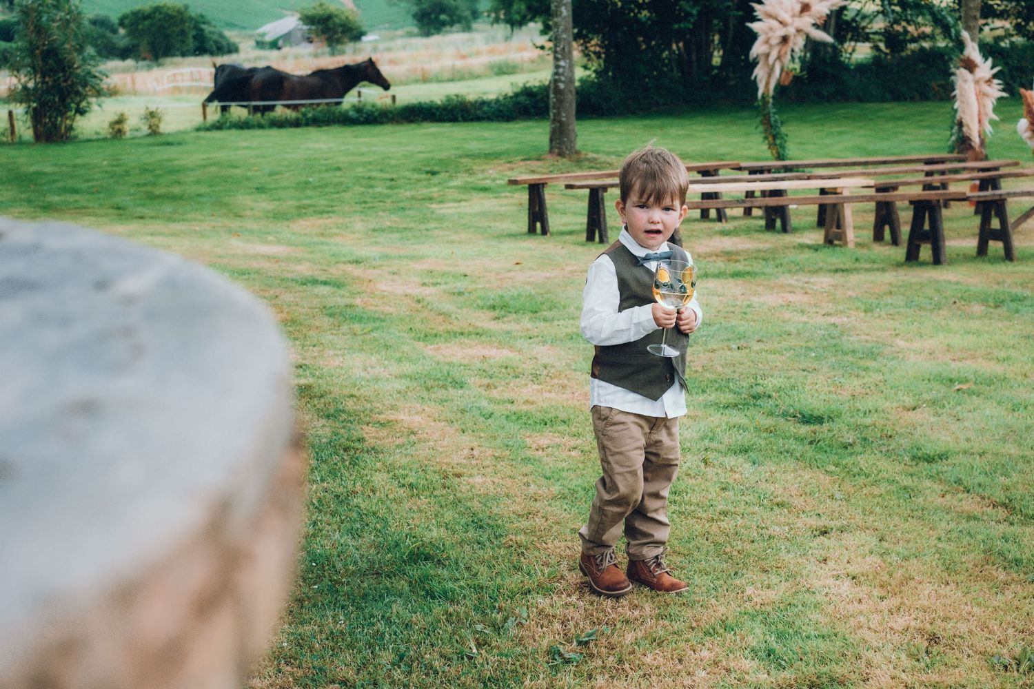Young ring bearer in vest stands alone in grassy outdoor wedding venue.