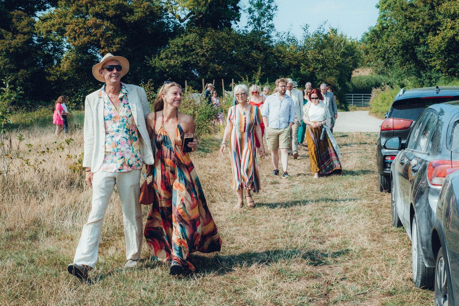 Guests mingling in an open field near parked cars at a rural outdoor gathering.