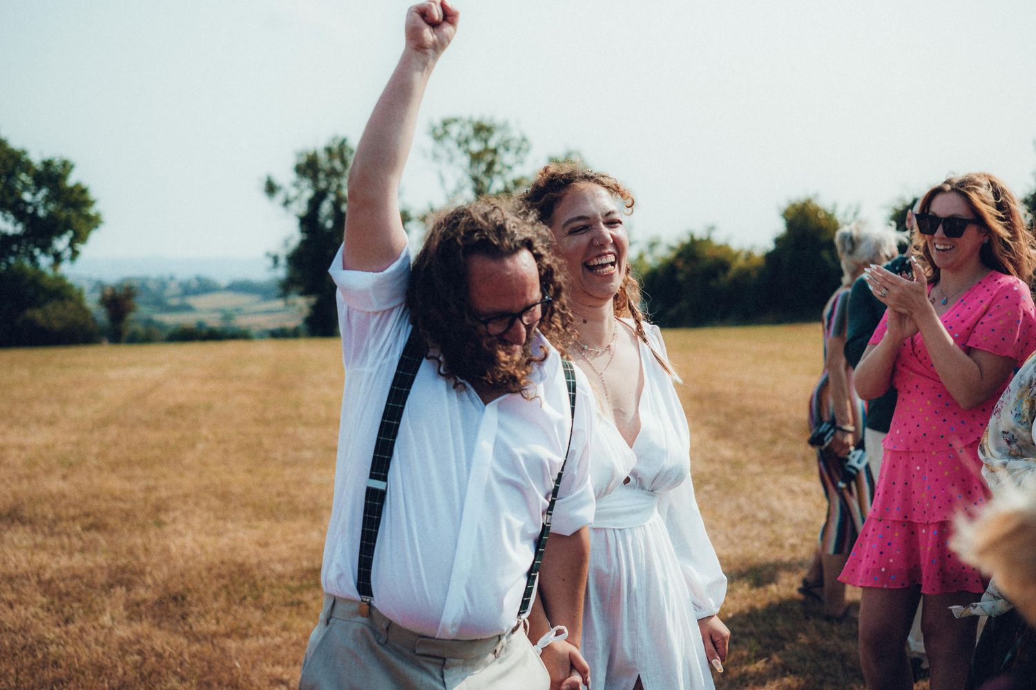 Wedding celebration outdoors with guests in white attire against scenic countryside backdrop.
