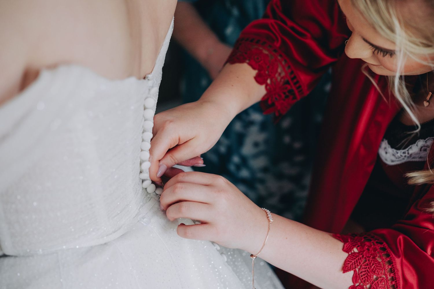 Red velvet sleeve and hands seen adjusting delicate lace detail on white wedding dress fabric.