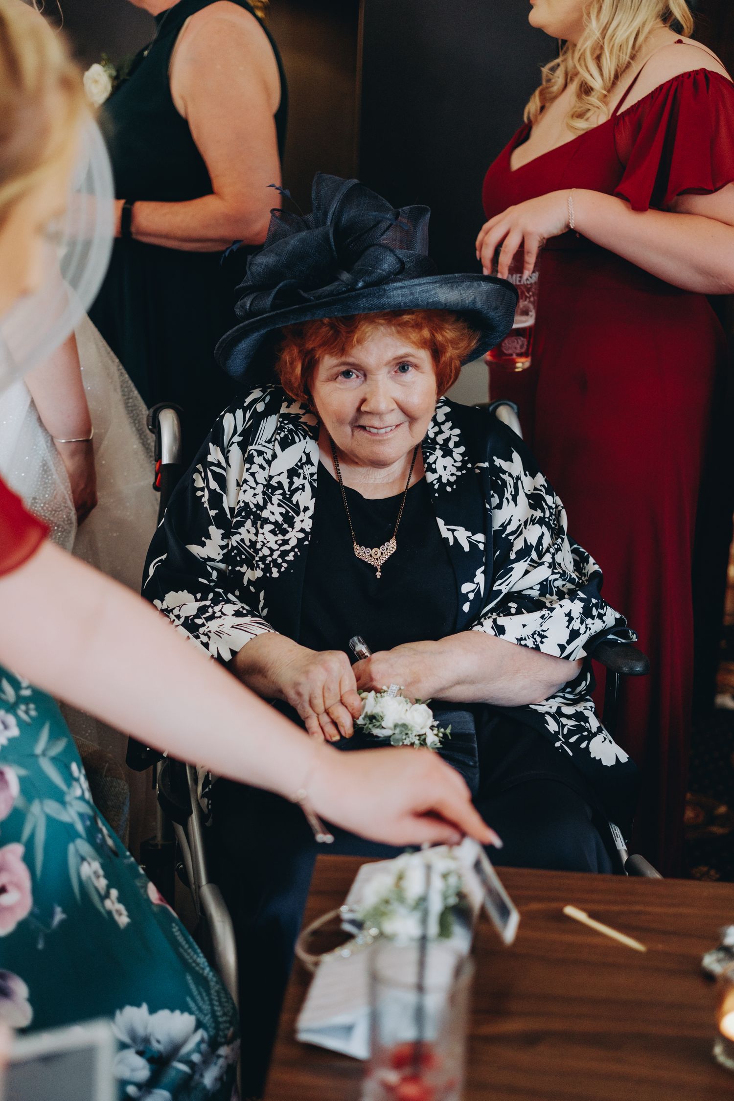 Woman in black and white floral dress and dark hat sits at table during wedding drinks reception.