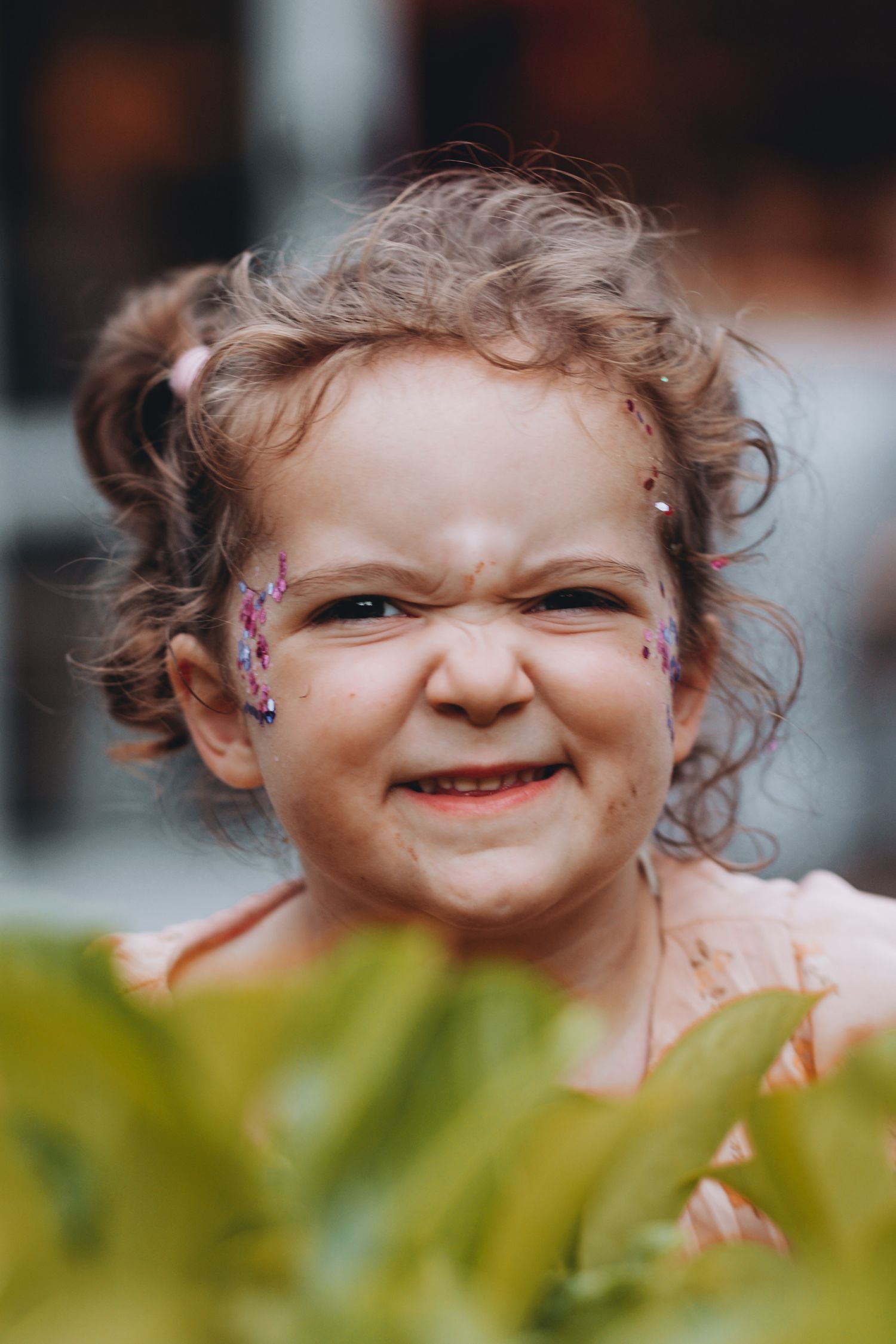 A joyful young child with curly hair and face decorations grins expressively among green foliage at wedding at Bartle Hall in Preston.