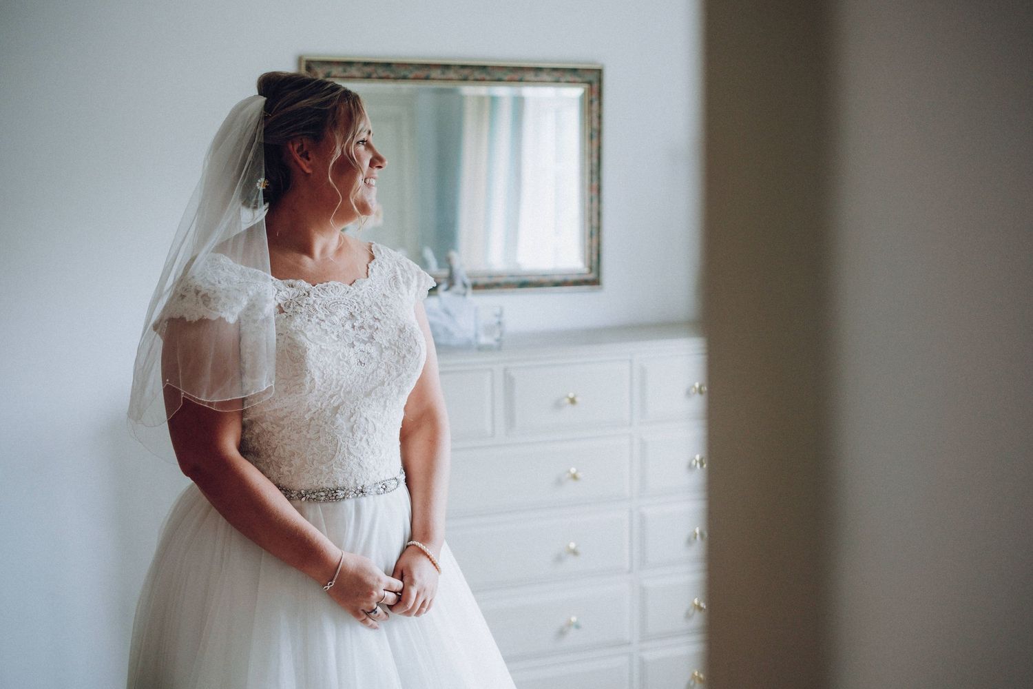 Side view of a bride in a classic white lace wedding dress standing by a dresser.