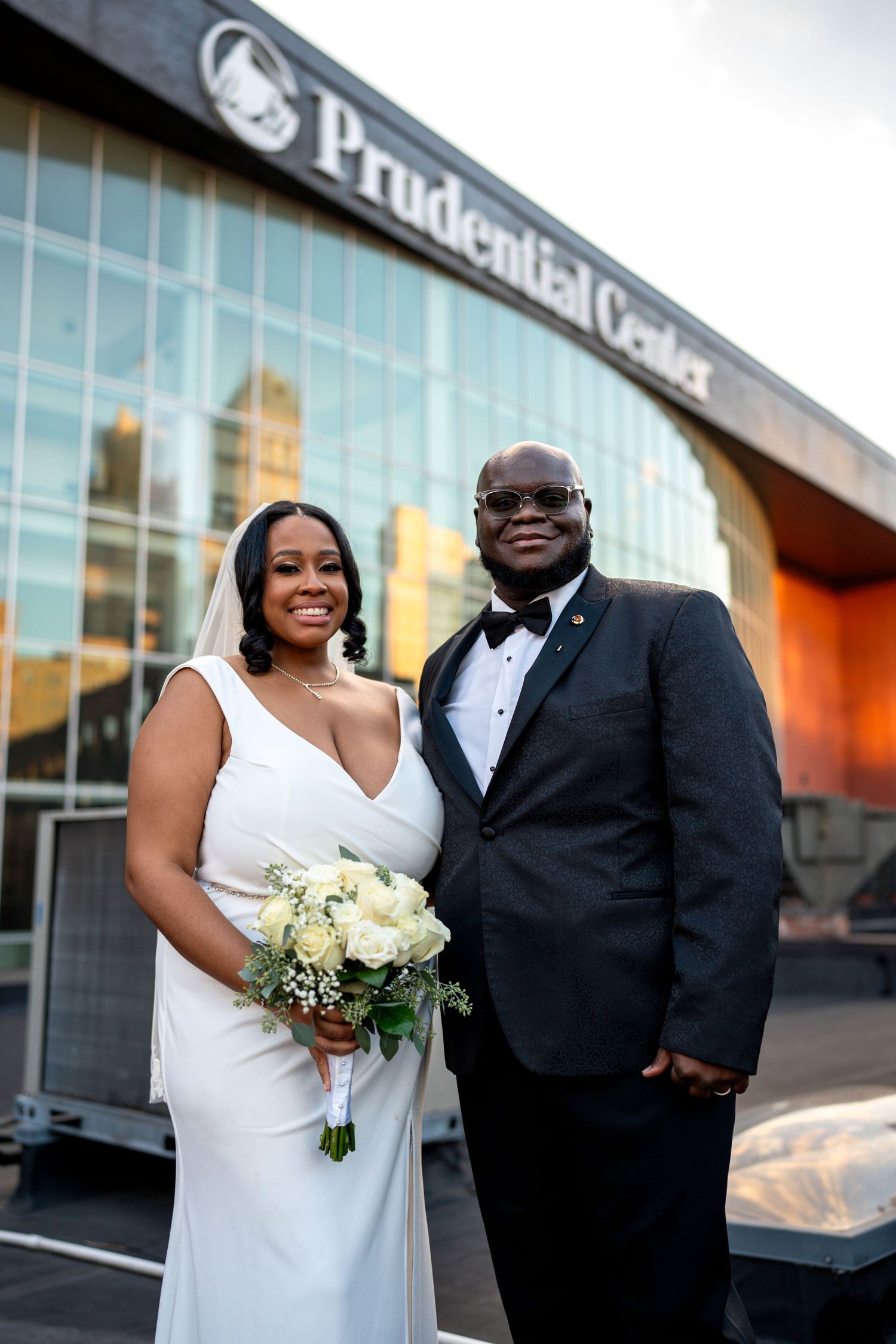 Elegant bride holding white bouquet stands with tuxedoed groom before Prudential Center.