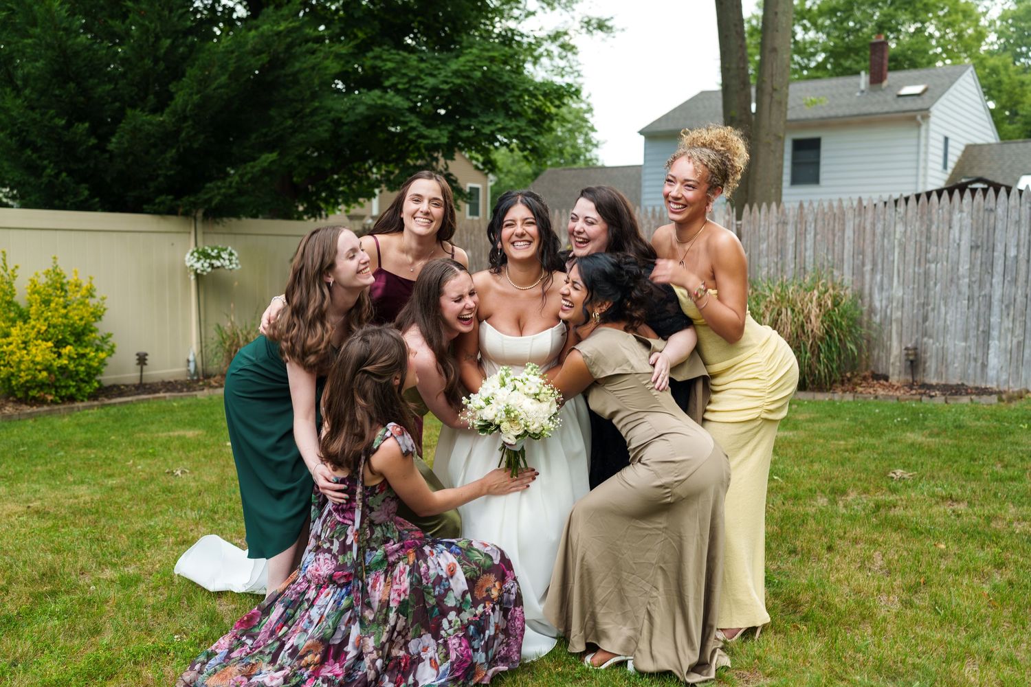 Bride in white gown surrounded by laughing bridesmaids in colorful dresses on green lawn.