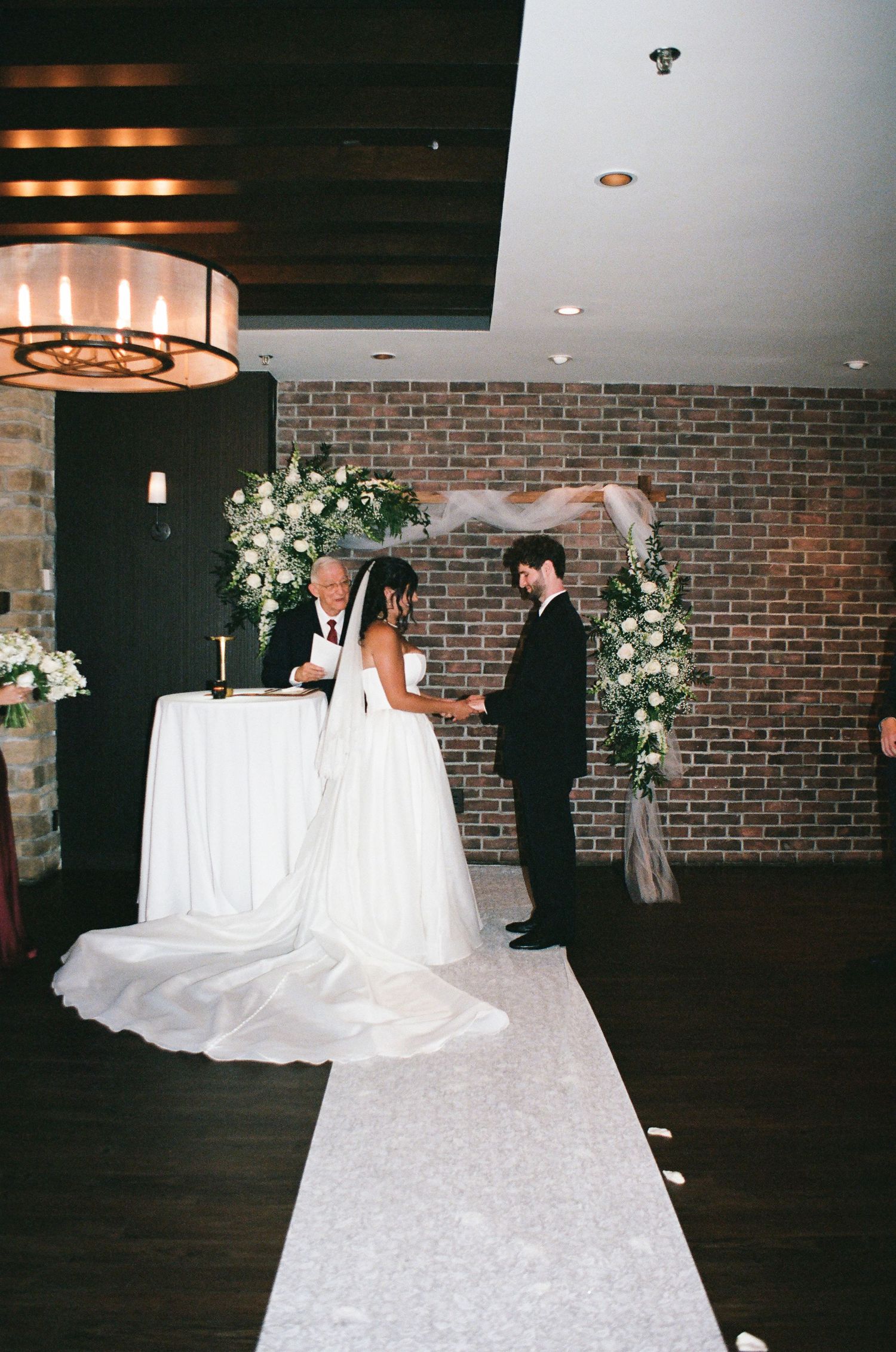 Bride and groom exchanging vows at indoor ceremony with brick wall and floral arch.