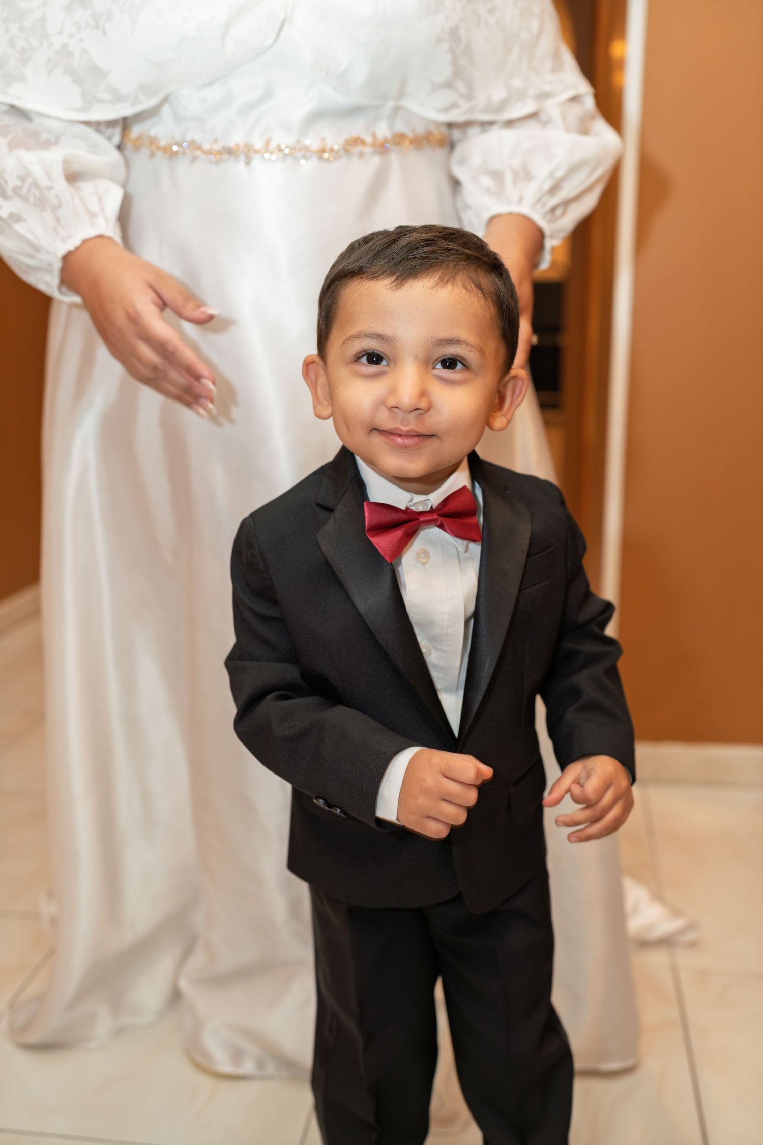 An adorable young ring bearer in a black tuxedo with red bow tie smiles at a wedding.