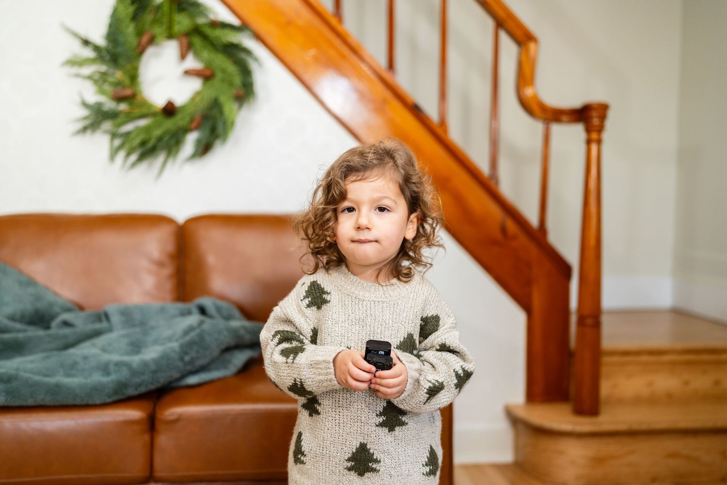 Young child in Christmas tree sweater holding a remote control near holiday decor.