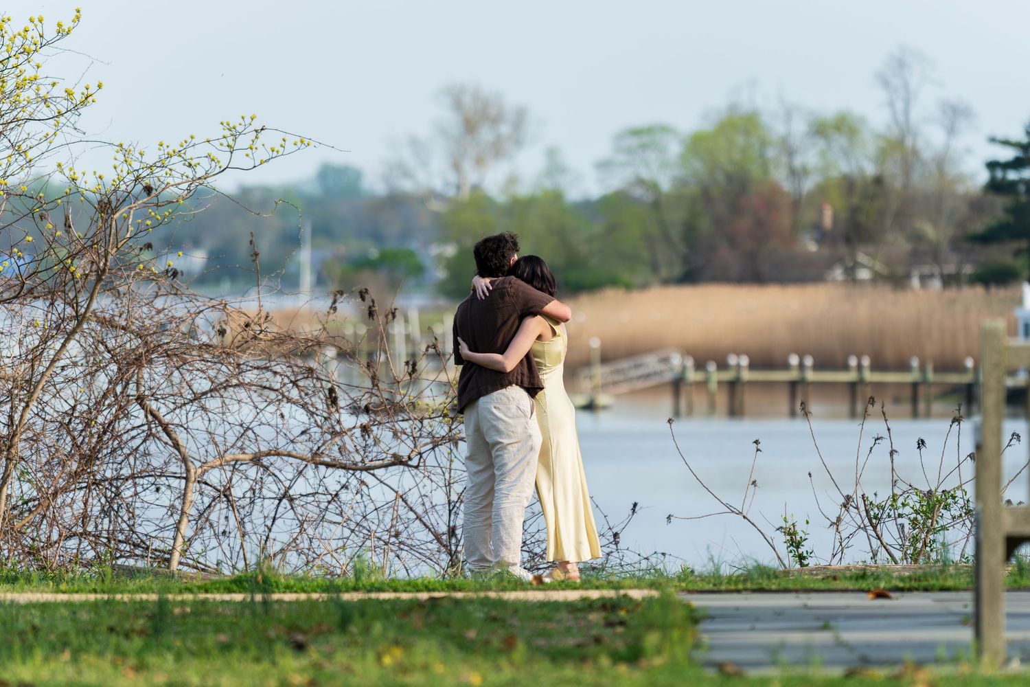 Couple embracing on a waterfront boardwalk surrounded by spring trees and calm water.
