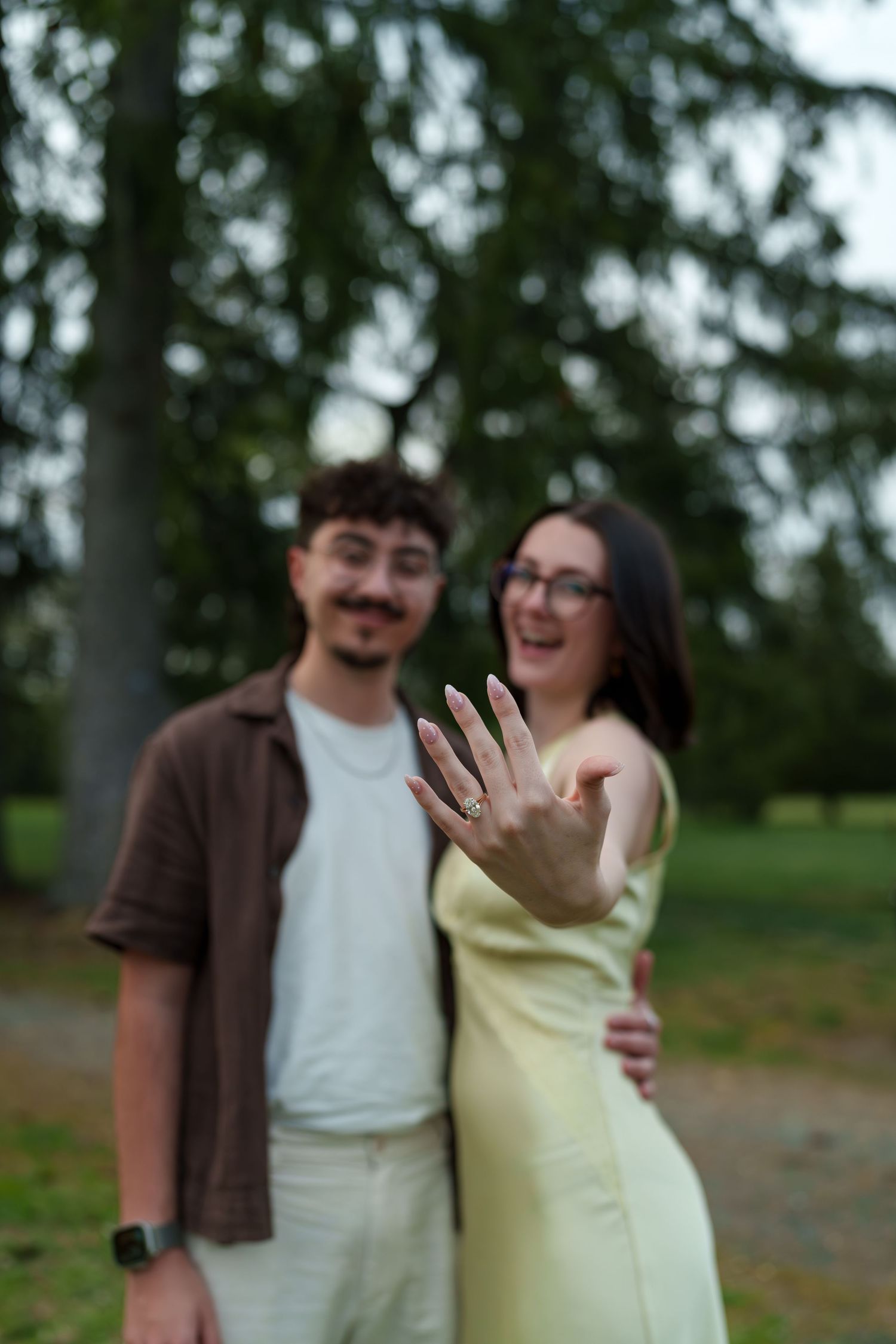 Excited newly engaged couple showing off engagement ring to camera in a green park setting.