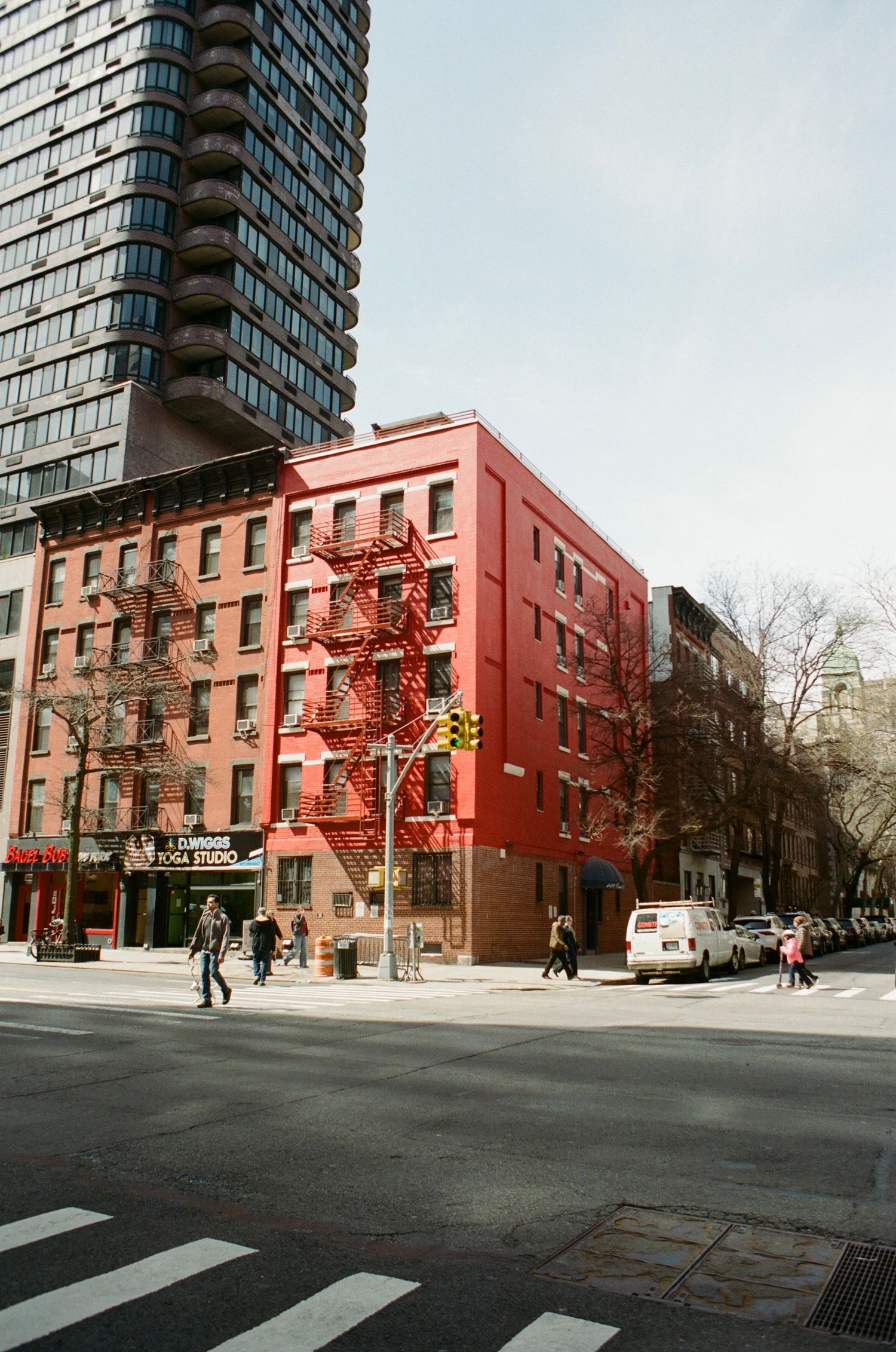 Pedestrians walk past a red brick building on a New York City street corner with a skyscraper behind.