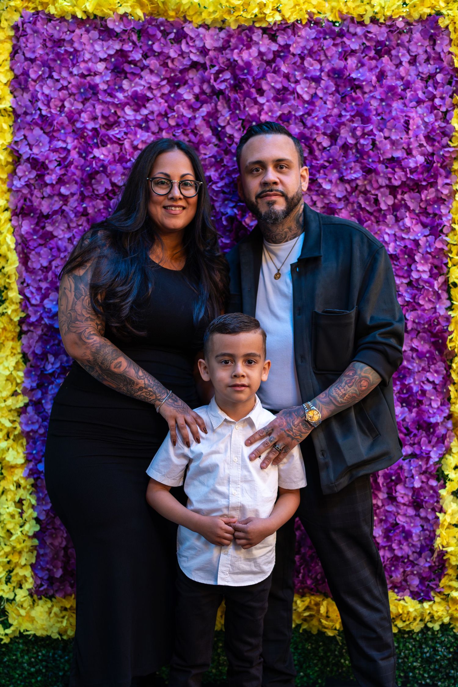 Family of three posing in front of purple and yellow flower wall backdrop.
