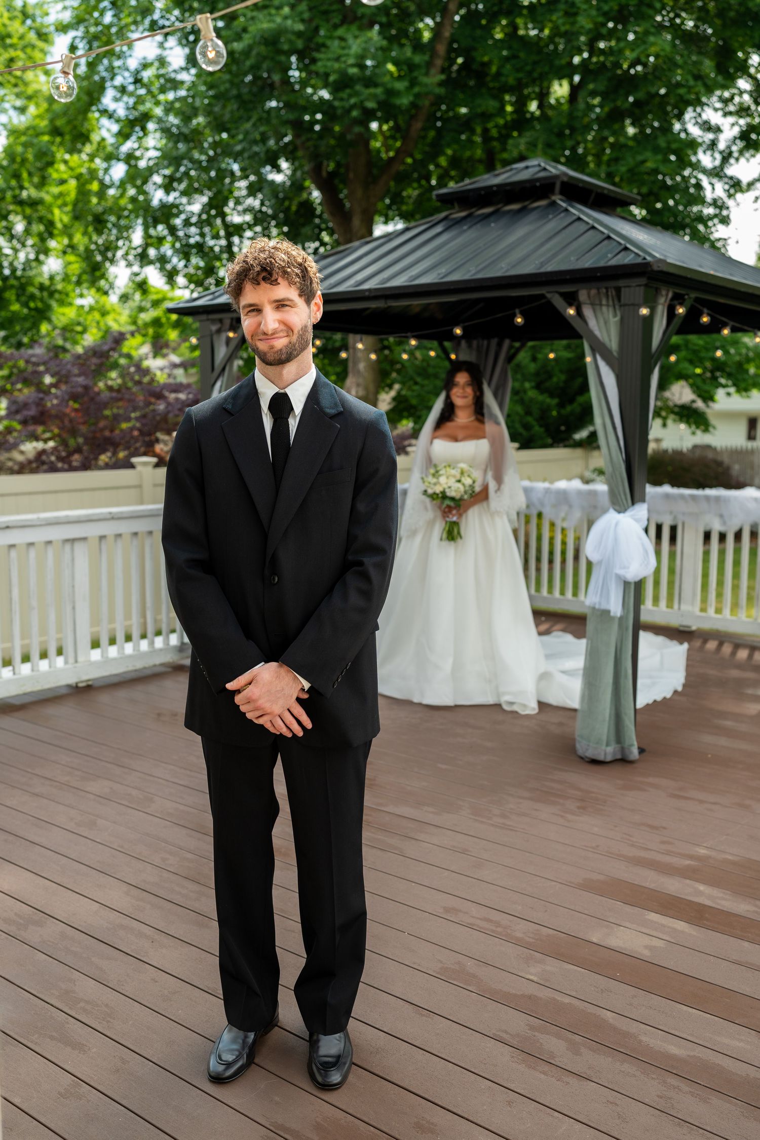 Groom in black suit smiling as bride in white gown approaches gazebo during outdoor first look.