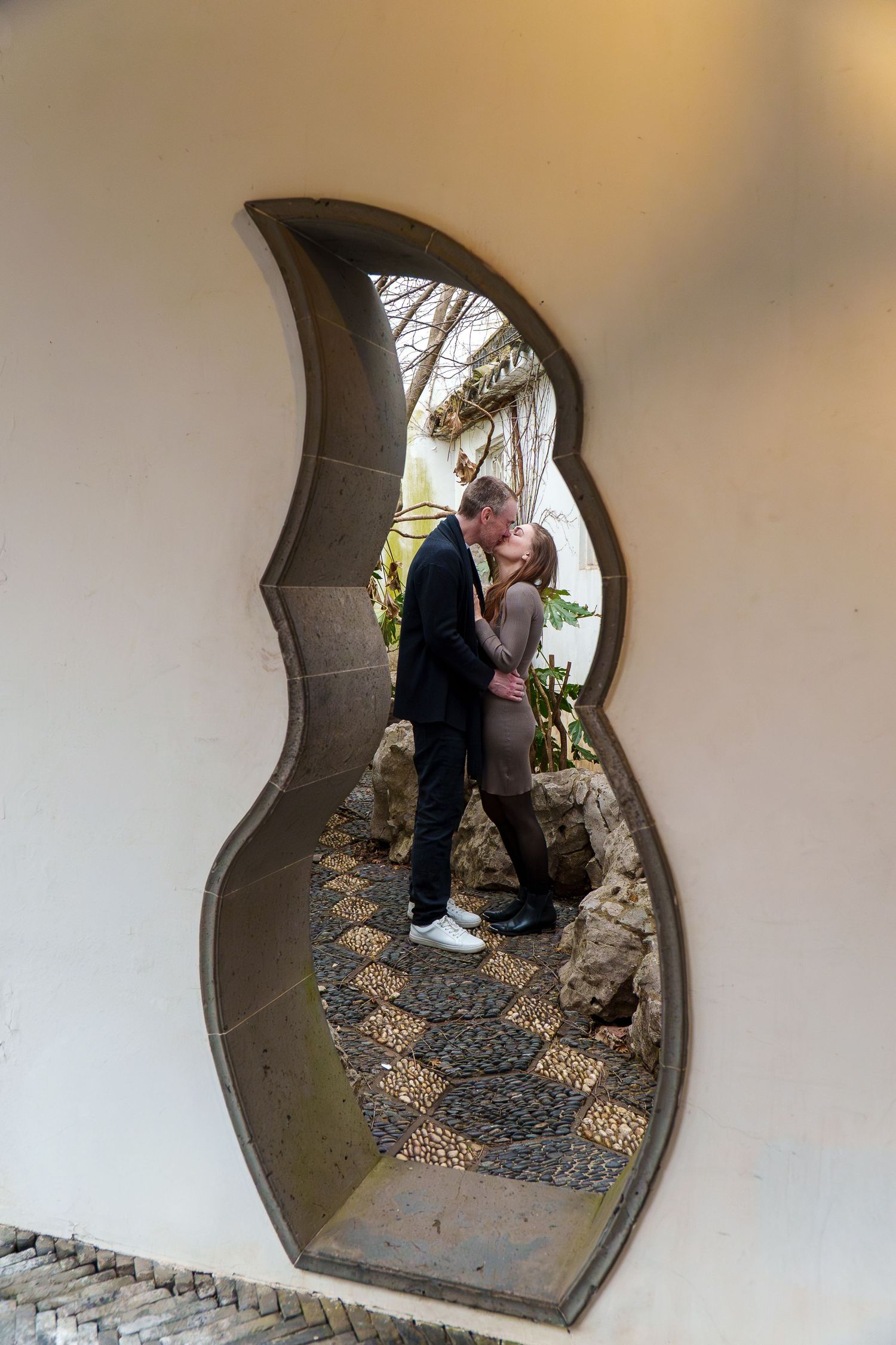 Couple sharing a kiss framed by a decorative flame-shaped moon gate opening in a Chinese garden.