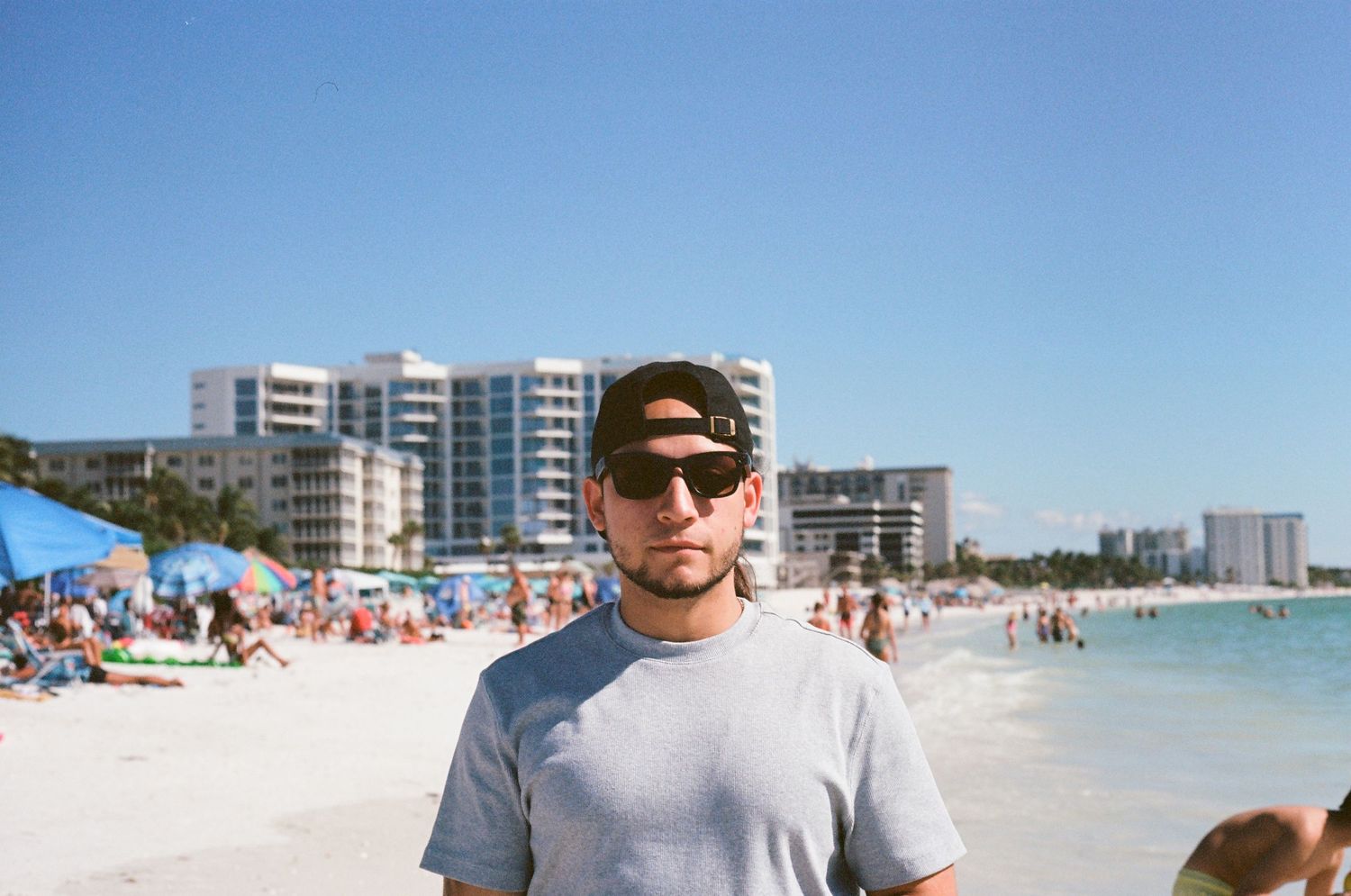Young man in cap and sunglasses standing on a crowded white sand beach with ocean behind him.