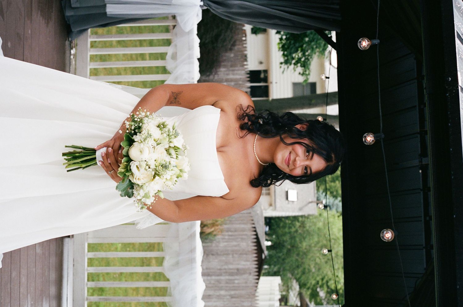 Smiling bride in white strapless wedding gown holding white floral bouquet outdoors.
