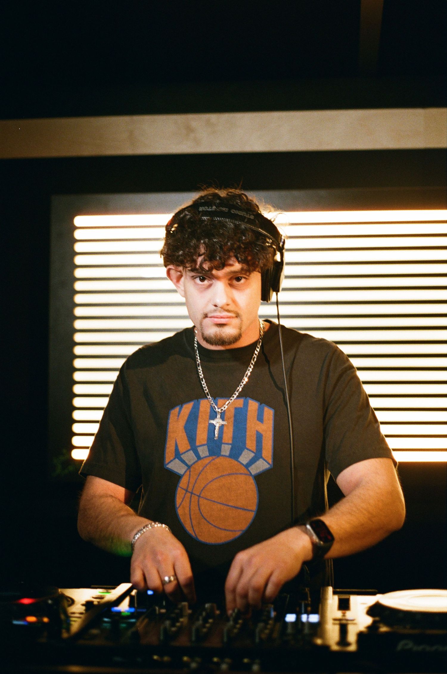 DJ wearing KITH Knicks shirt mixing music with headphones in dimly lit studio.