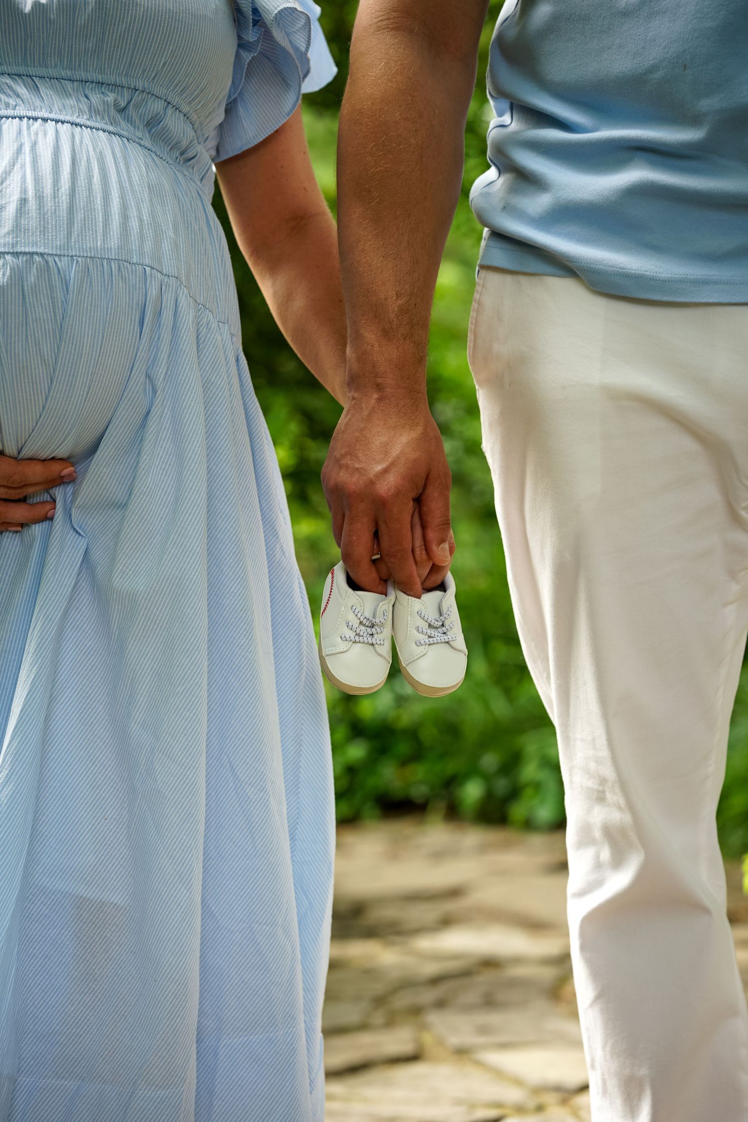 Expectant couple holding tiny white baby shoes while standing on stone path in green garden.