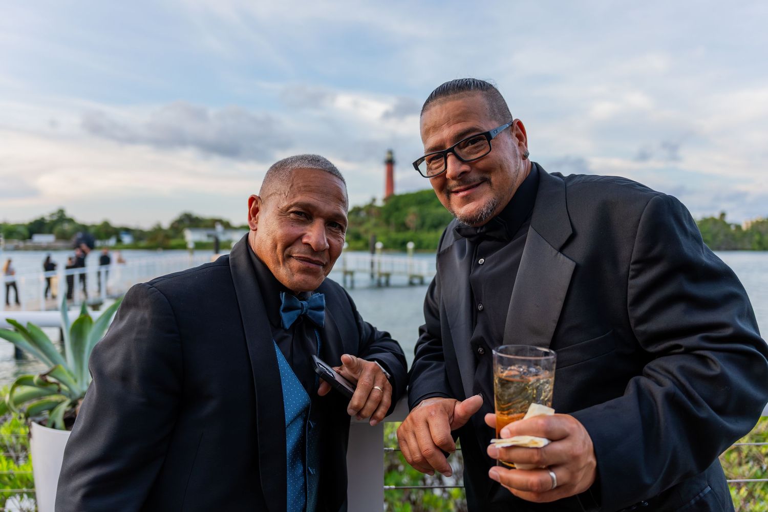 Two men in suits smile outdoors at a waterfront event, one holding a drink and cigar.