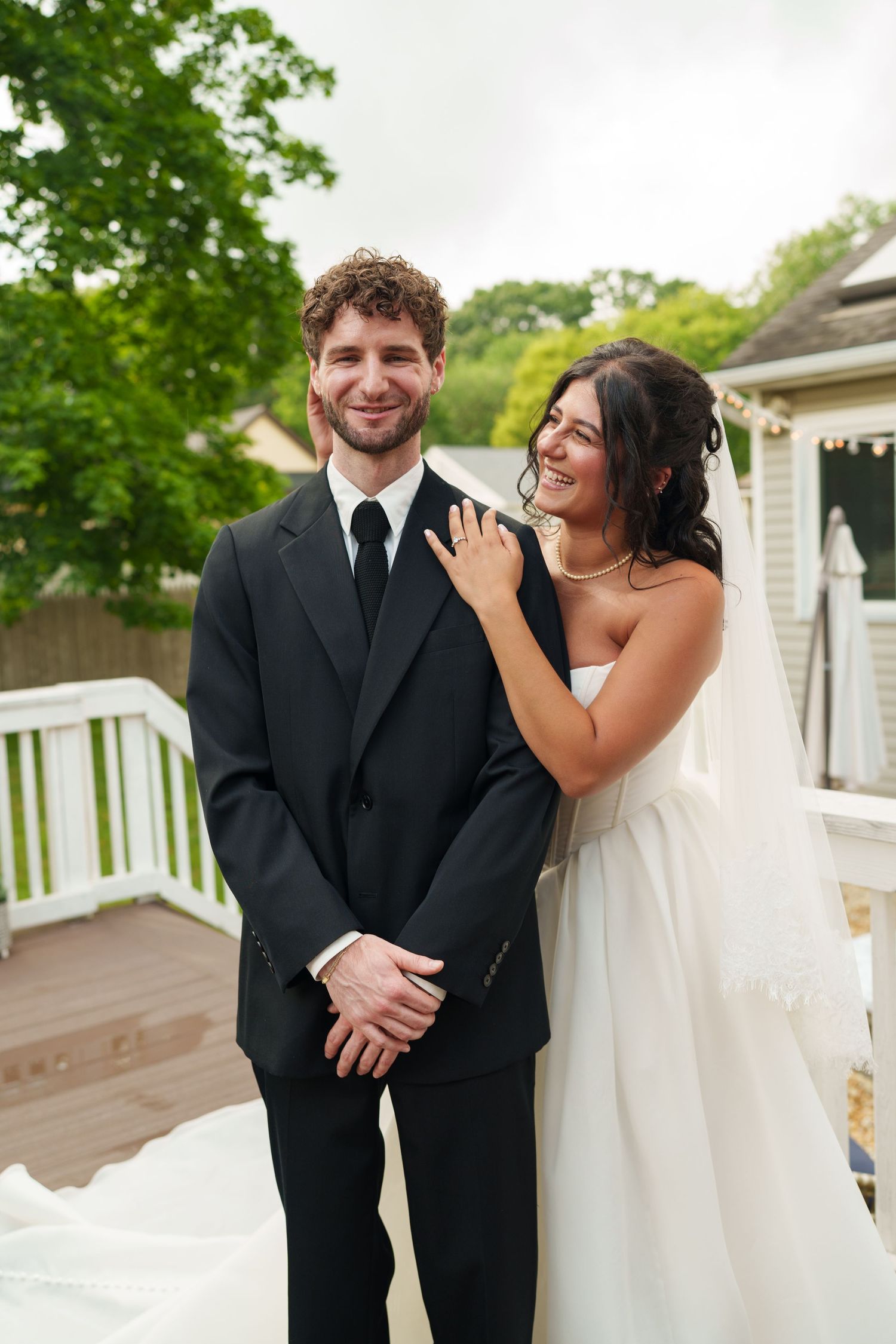 Smiling groom in black suit stands with bride in white gown on outdoor deck at wedding.