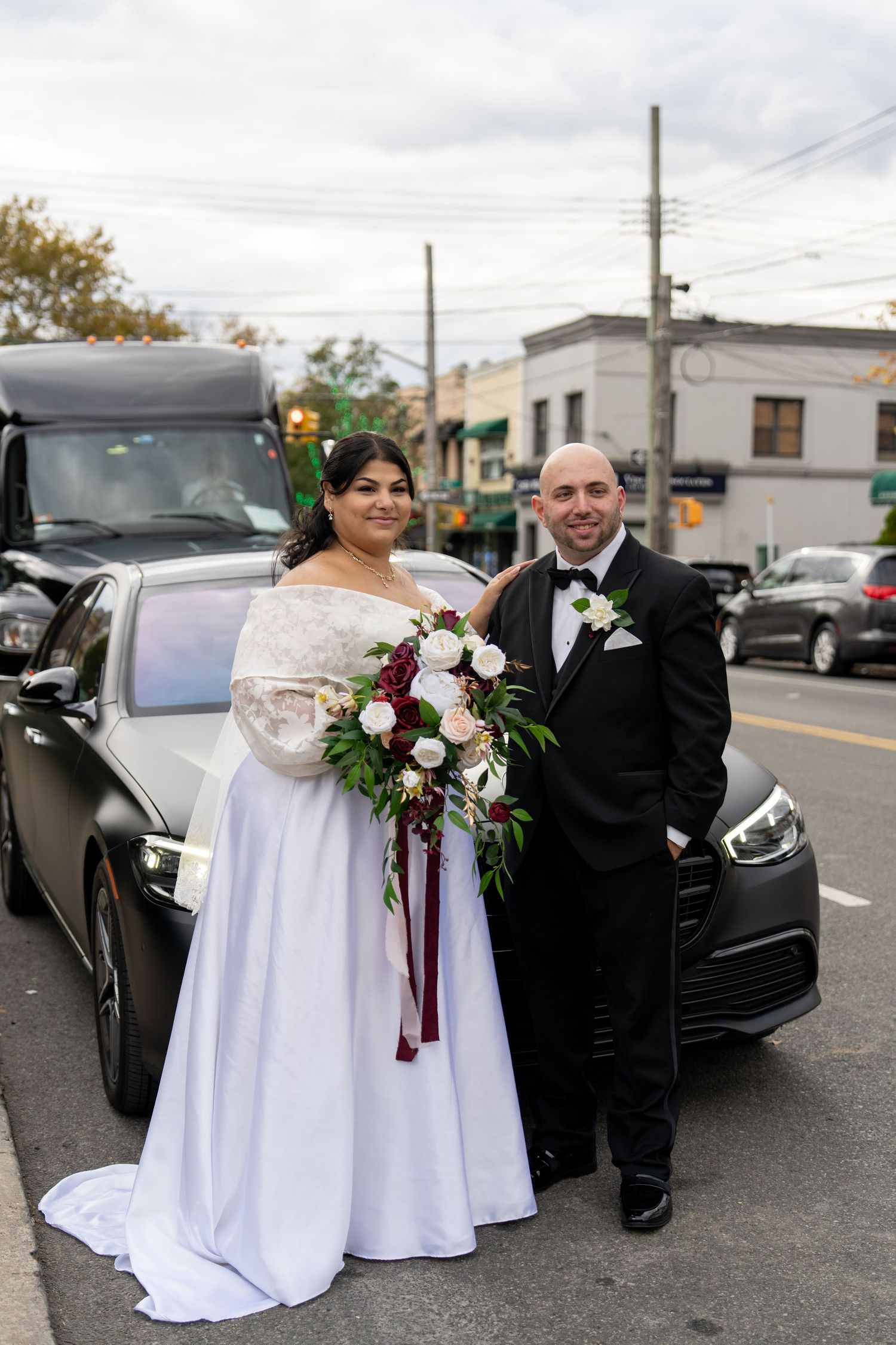 A bride and groom pose together in front of a black car on their wedding day.