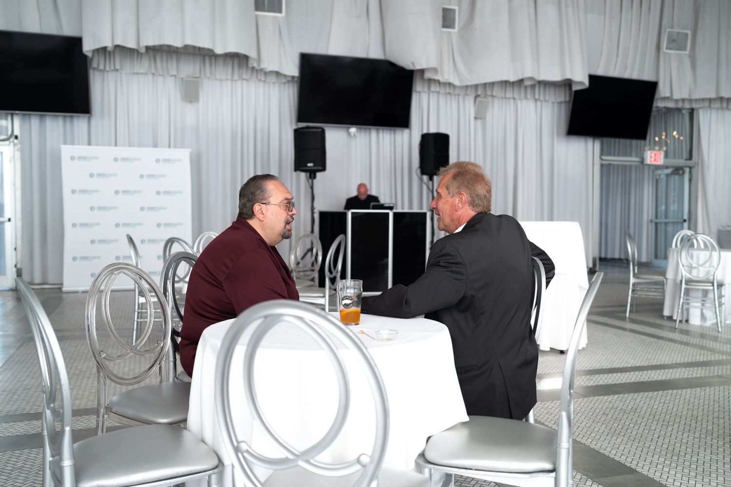 Two men having a conversation at a high-top table in a white decorated event venue with DJ.